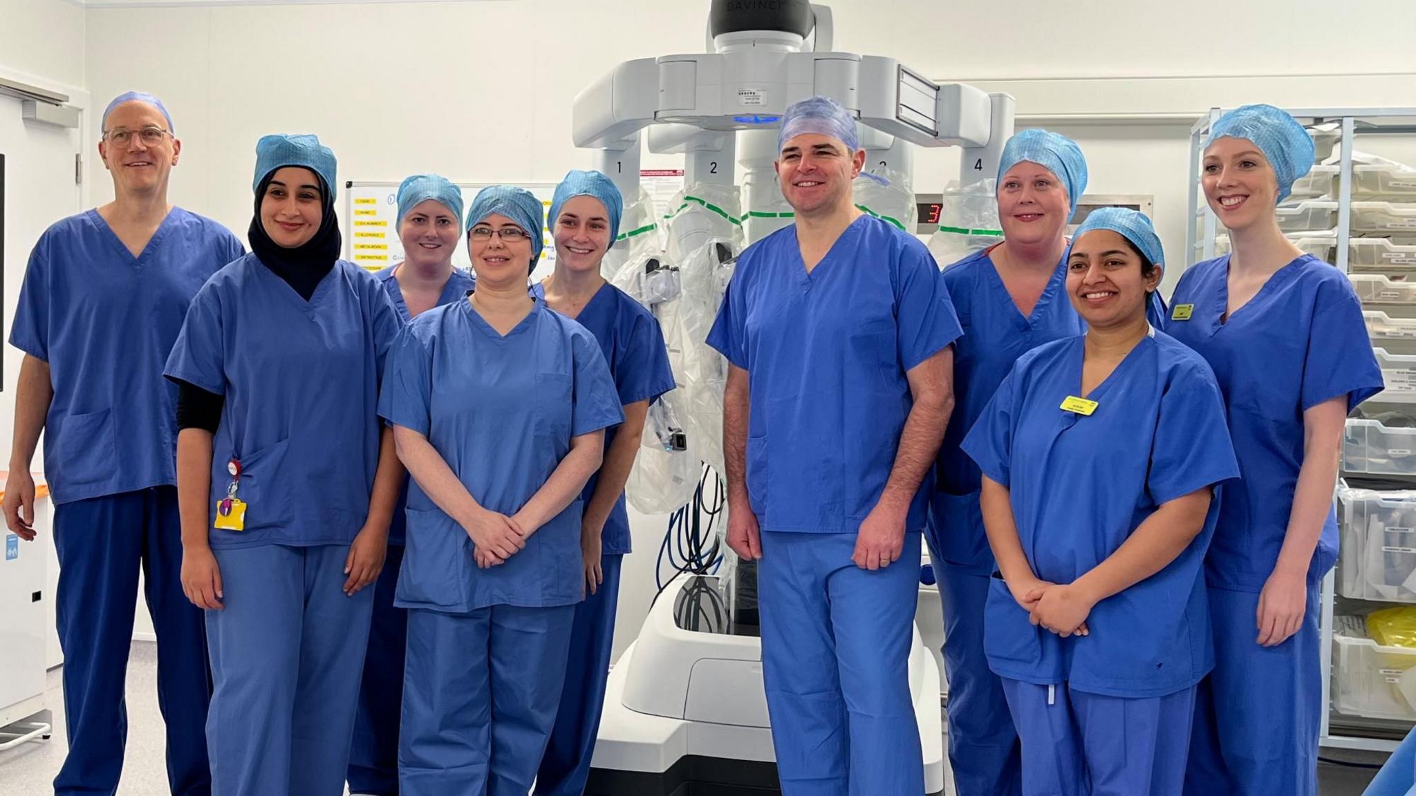 A photo of the full surgical team standing in front of the robot in the operating theatre. There are nine people, all wearing blue surgical scrubs and caps. 