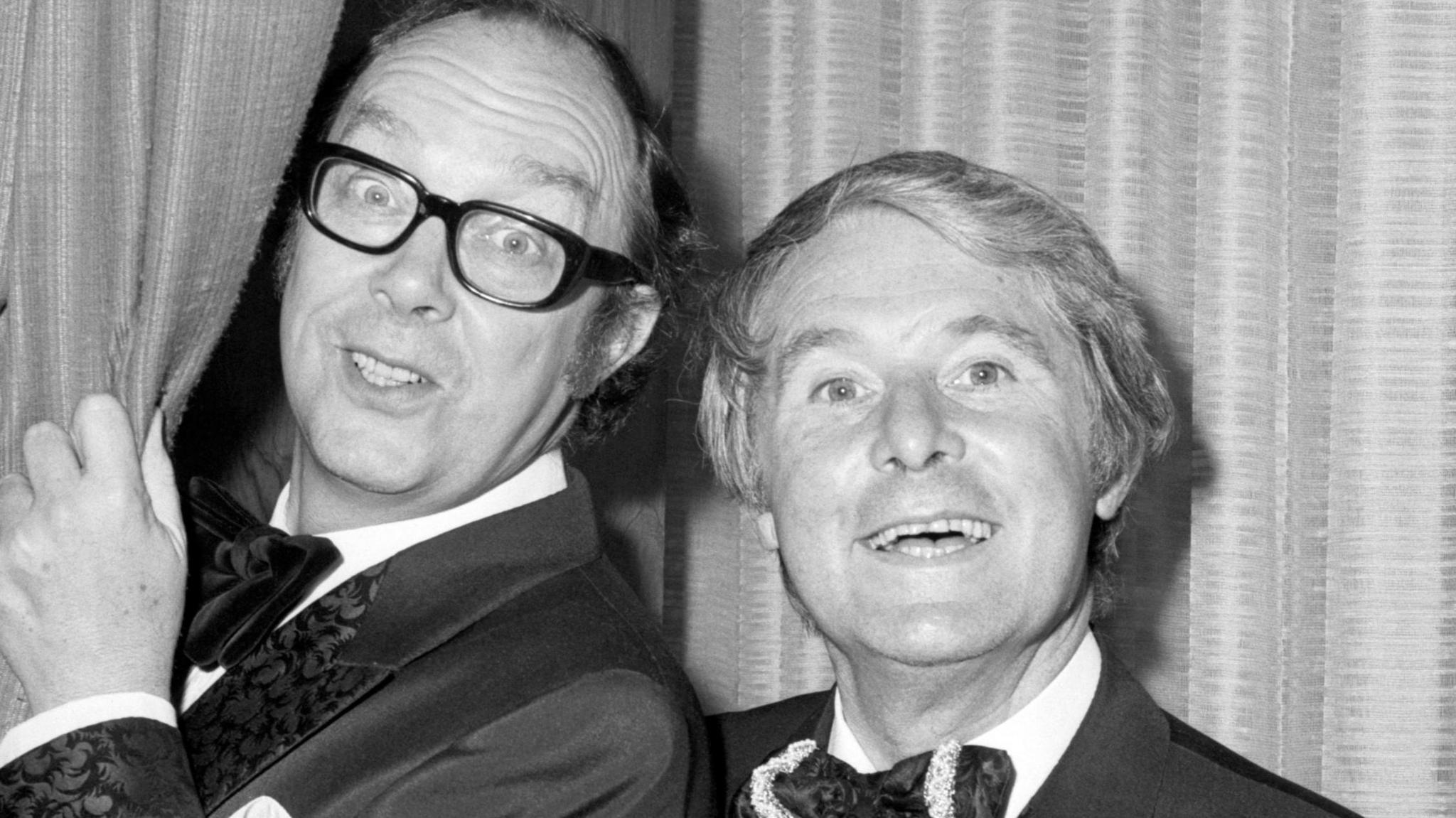 Eric Morecambe and Ernie Wise in black and white on the set of one of their tv shows. Both men wear suits and bow ties, Mr Morecambe is poking his head out of the curtains behind him and wears thick black framed glasses. Mr Wise is smiling next to him at the camera.