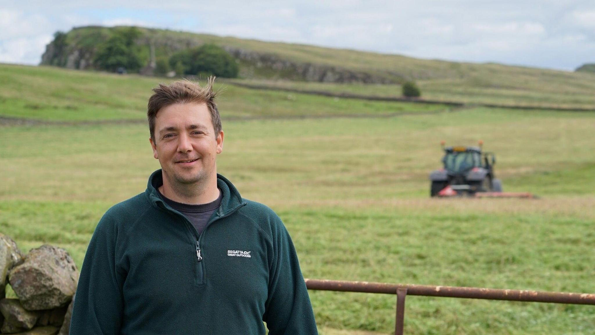 Kit Acton smiling into the camera. He has short, brown hair which is sticking up and is wearing a green fleece. He is standing in front of a green field with a tractor out of focus in the background.