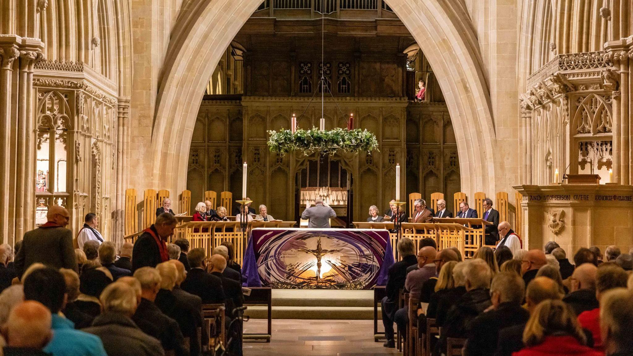 People attending a carol service inside Wells Cathedral. The pews are all filled with people and the choir is singing by the altar.