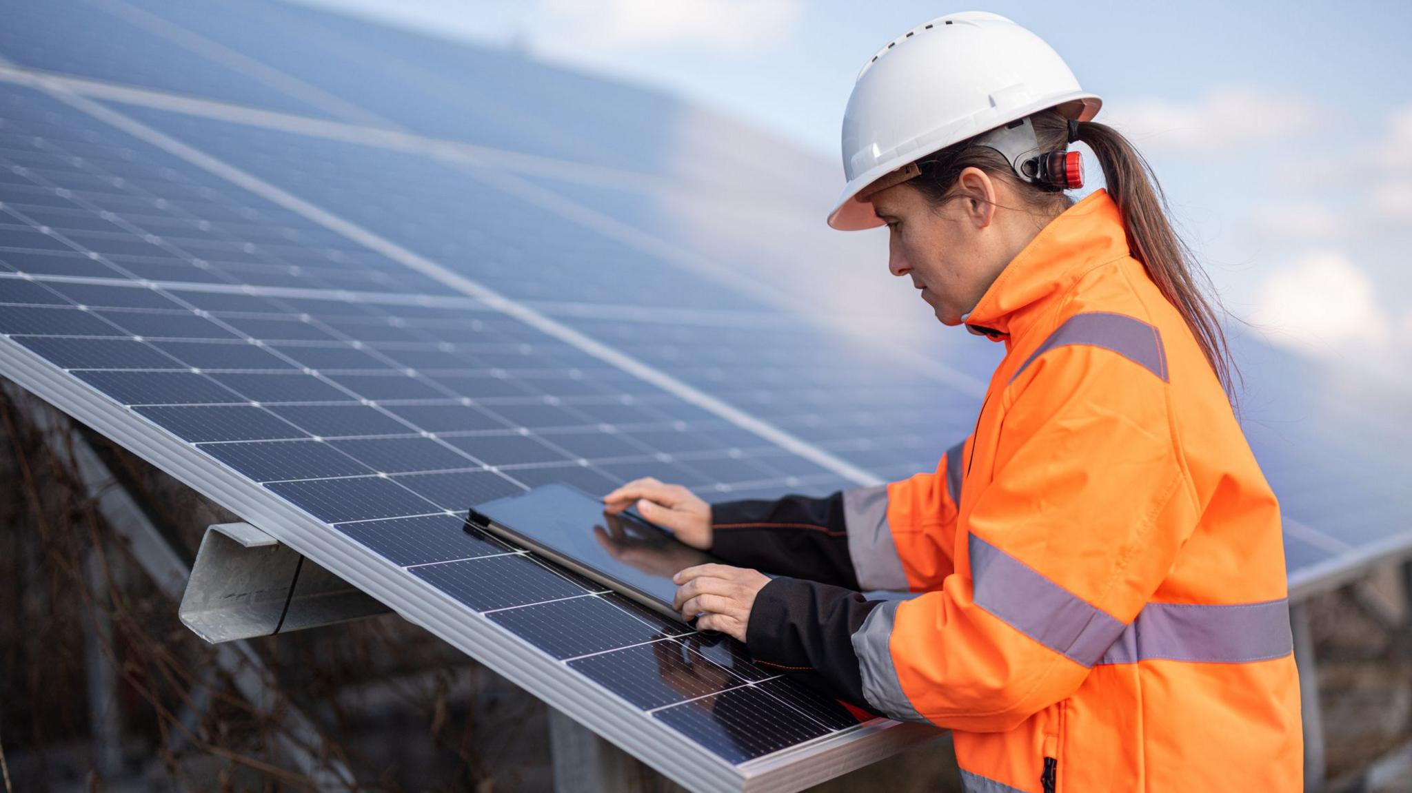 A woman in an orange high-vis jacket uses a tablet computer resting on a large solar panel