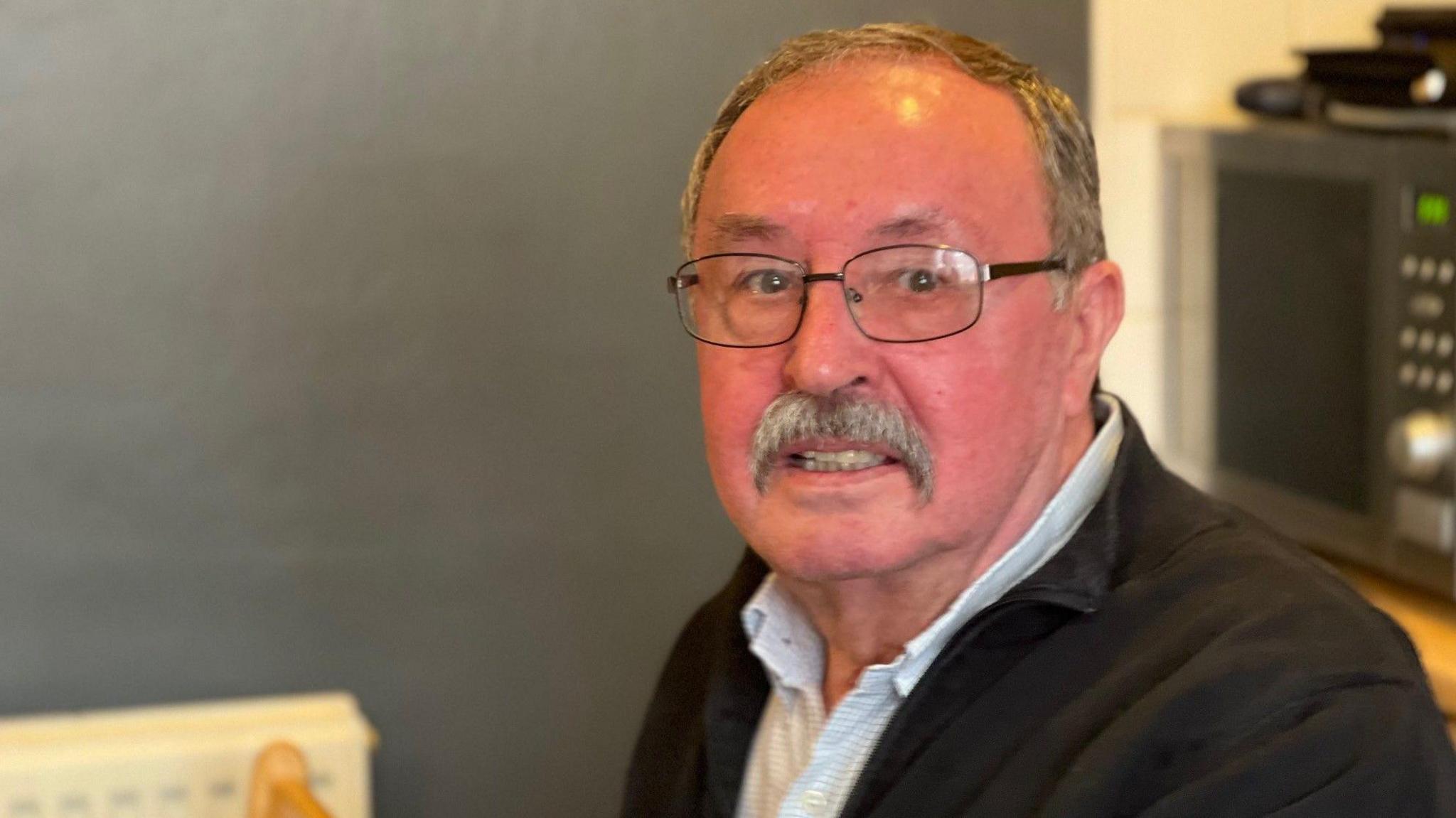 A man wearing glasses smiles at the camera. He is wearing a blue shirt and black jacket.