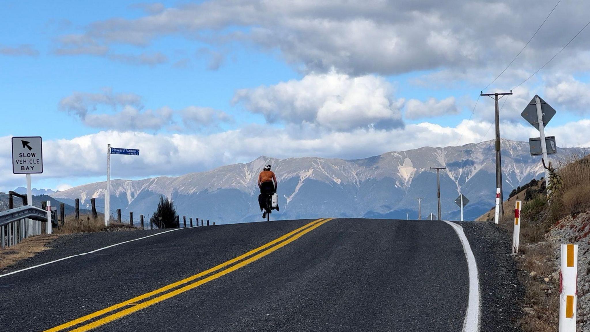 A picture of a long road in New Zealand. There is a cyclist travelling on the left side of the road. There is a number of mountains and low clouds in the distance.
