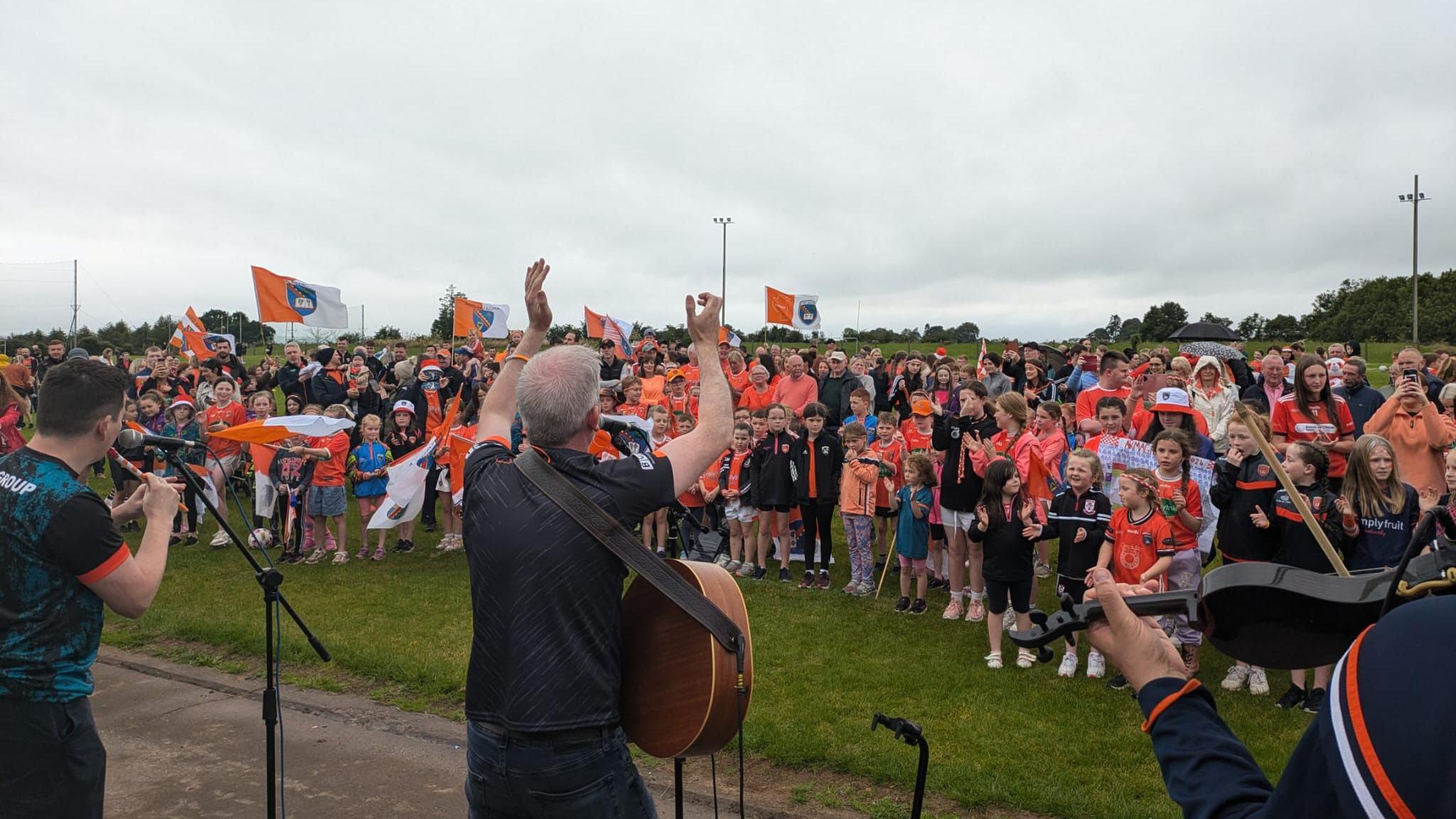 Man standing with a guitar with his back to the camera, clapping his hands above his head. He is facing a crowd standing on a GAA pitch.