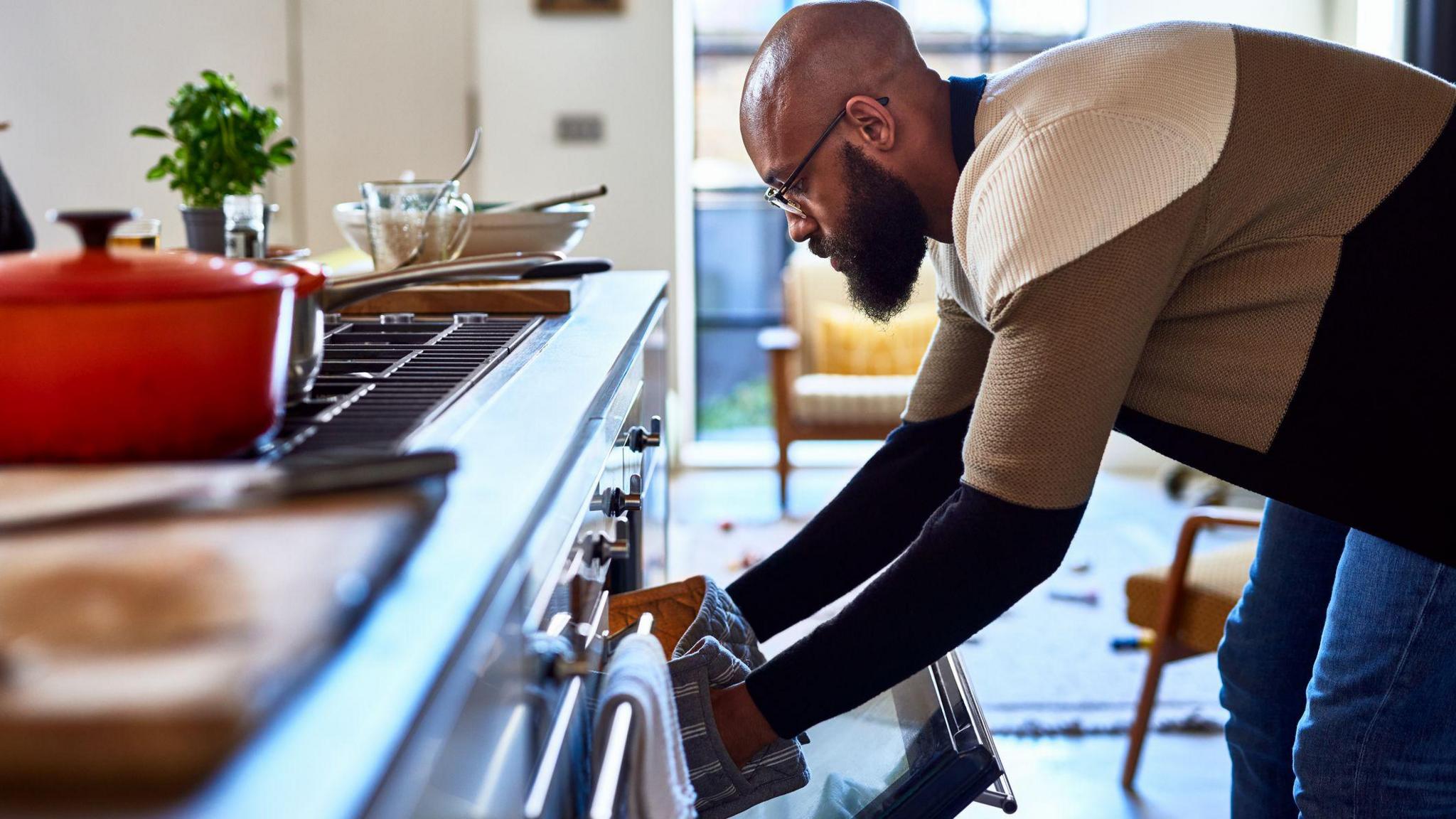 Man with his hands in oven gloves bends down to get something out of the oven, with the work surface and hob in front of him in the kitchen