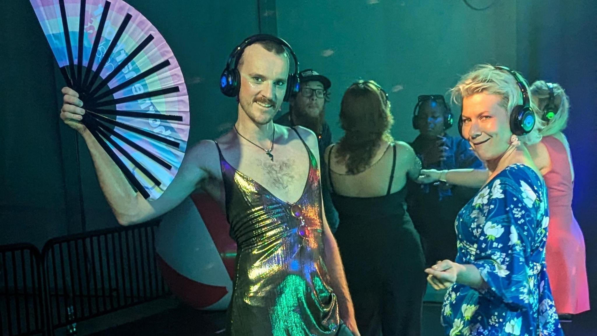 Two people are on a dance floor wearing headphones. One of them is holding a fan and they are both smiling. There are several other people in the background.