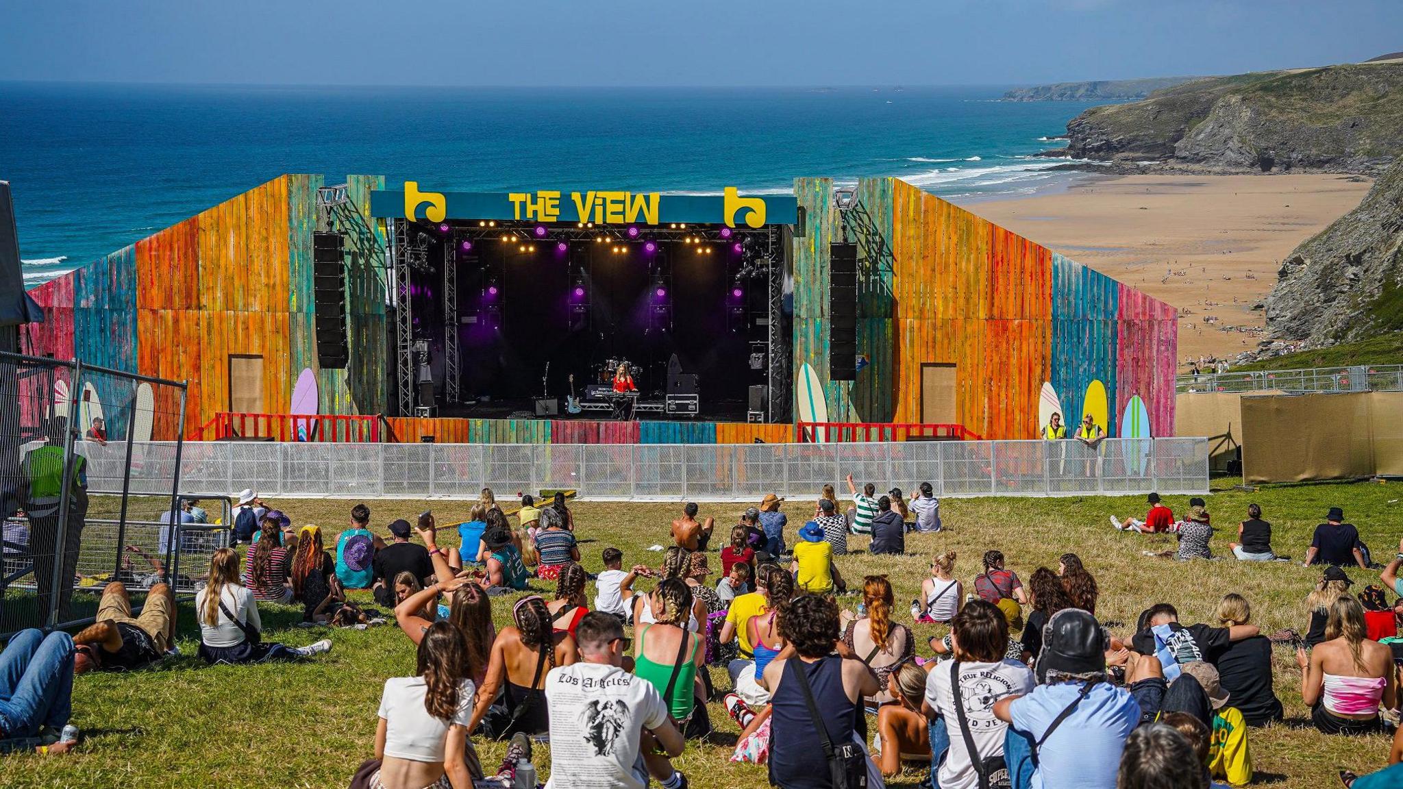 A colourful outdoor stage labelled 'THE VIEW' set up on a grassy area overlooking a beach and the ocean. The stage features vibrant multi-coloured panels and purple lighting. A small crowd sits on the grass watching a performance, with cliffs and the shoreline visible in the background.