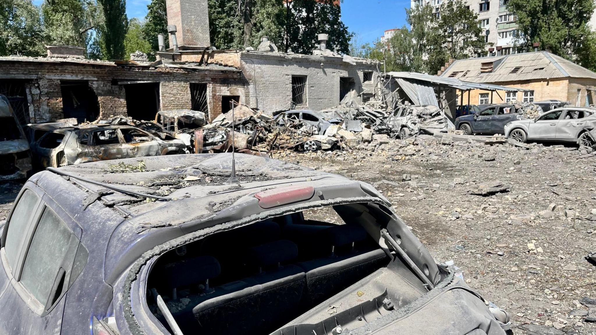 A line of burnt out cars covered in ash and debris, with buildings behind it that are heavily damaged