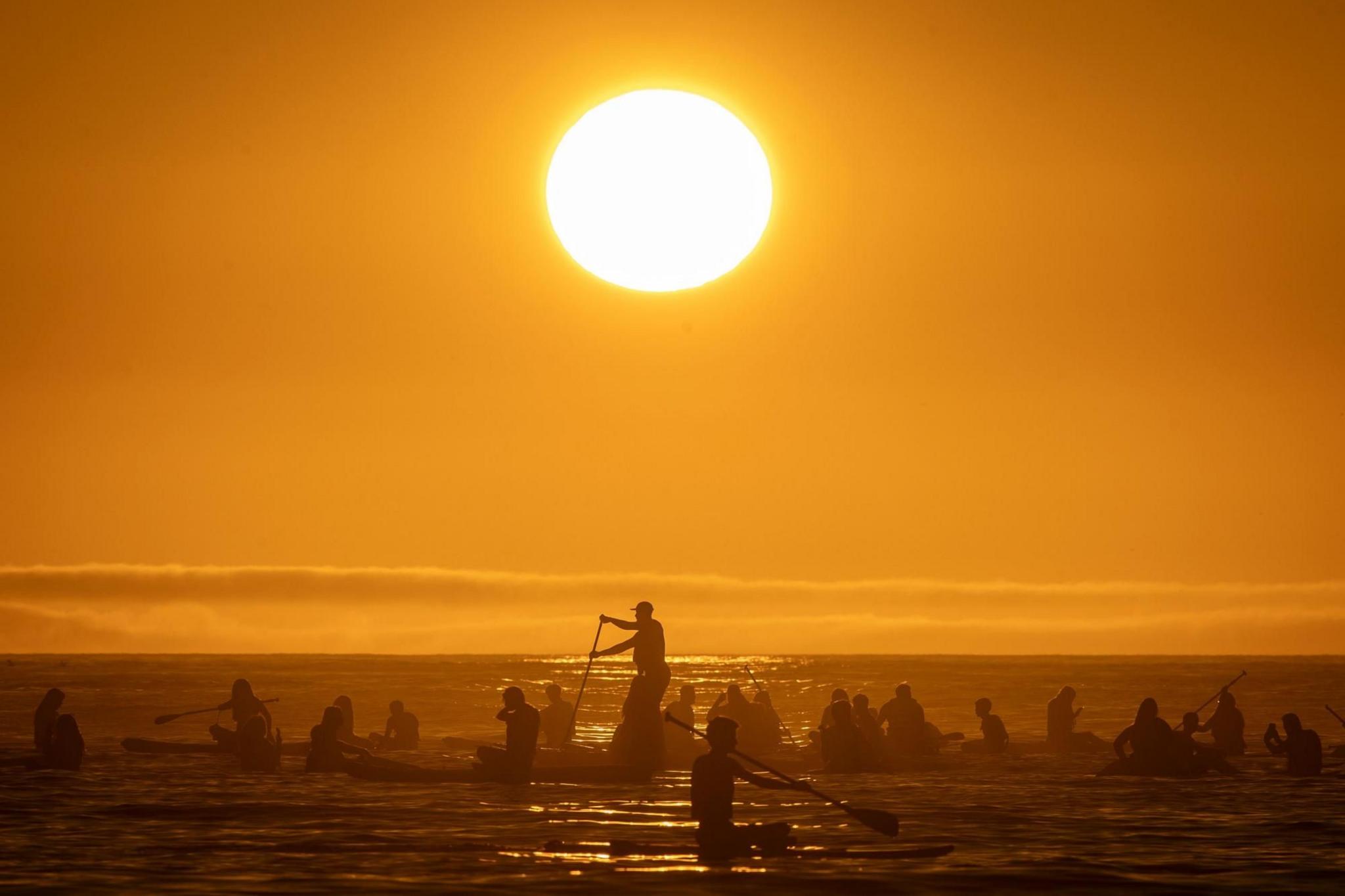 People sitting or standing on paddle boards in the ocean with the sun shining overhead in a bright yellow sky