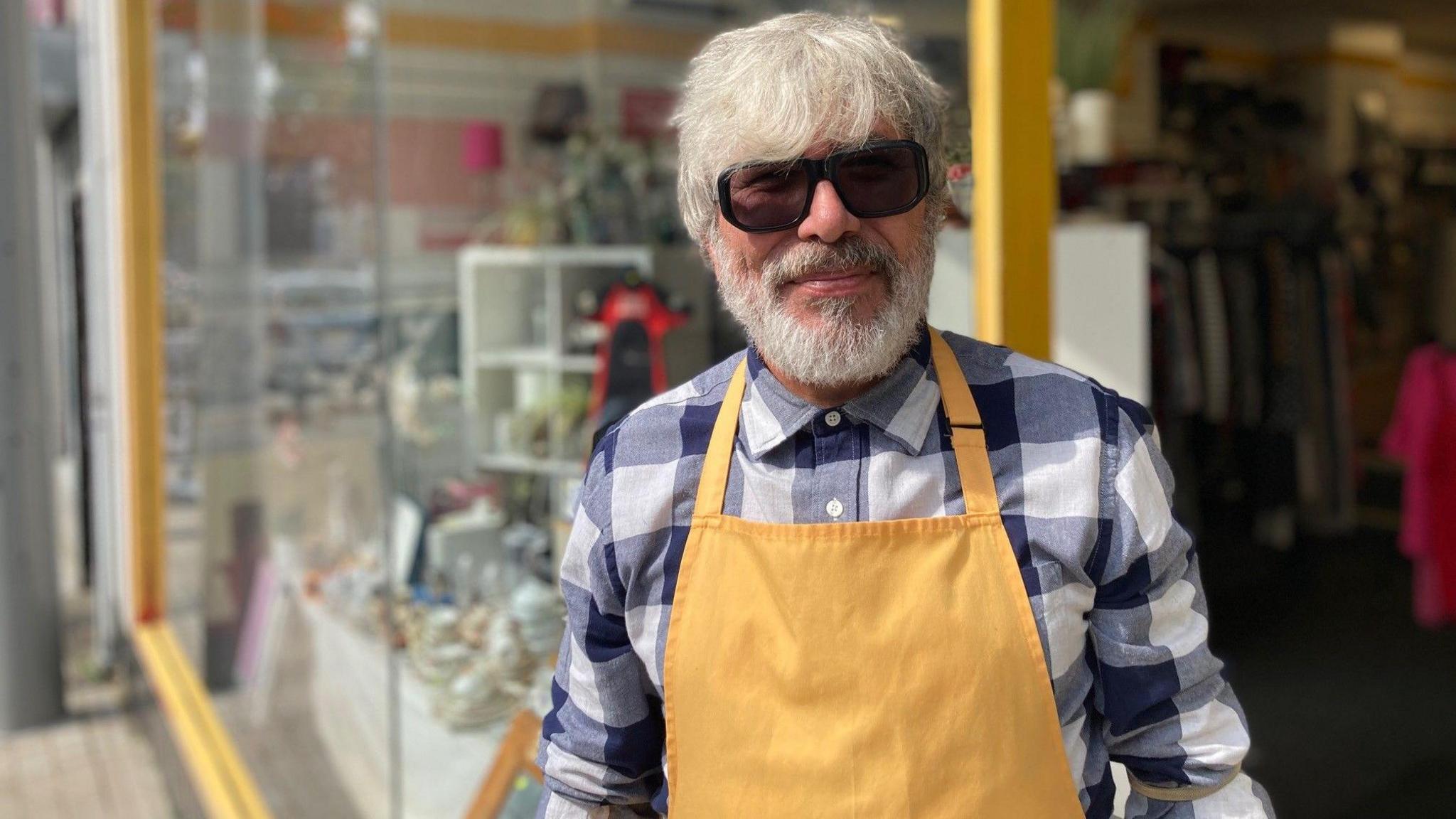 A man stands outside a shop. He is wearing a purple and white chequered shirt and a yellow apron. He is also wearing sunglasses.