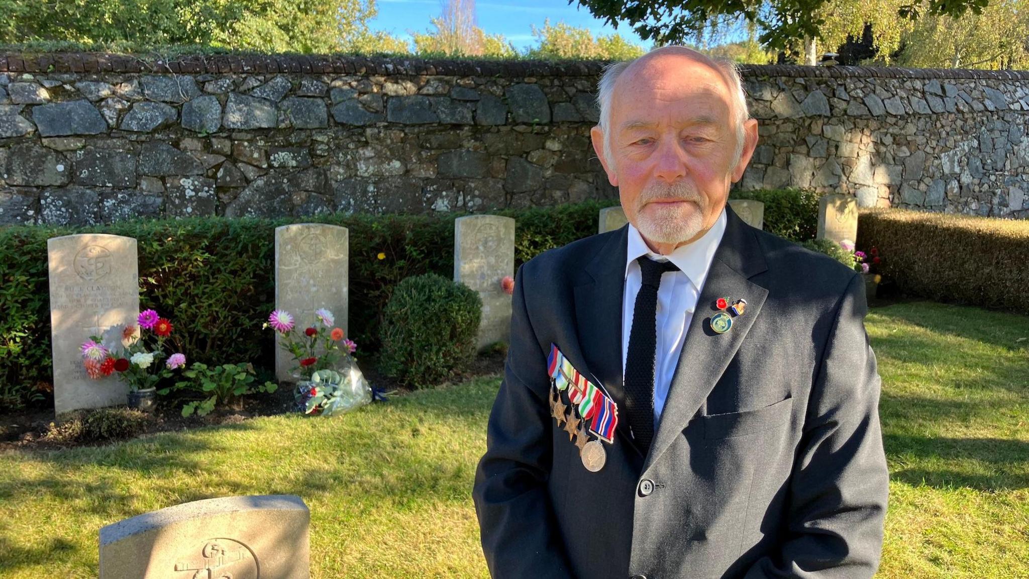 A man with receding grey hair and a well-kept grey beard in a blazer with medals on the lapel stands in front of a line of neatly-kept graves with flowers and wreaths in front.