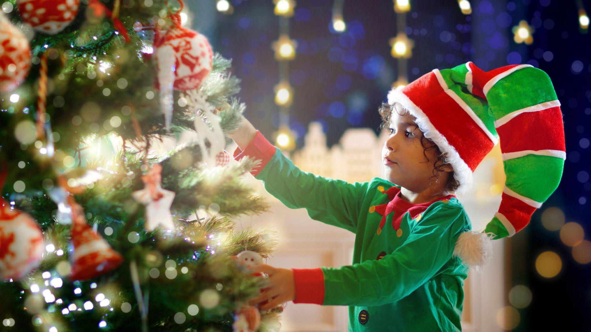 Boy dressed as an elf putting decorations on Christmas tree. 