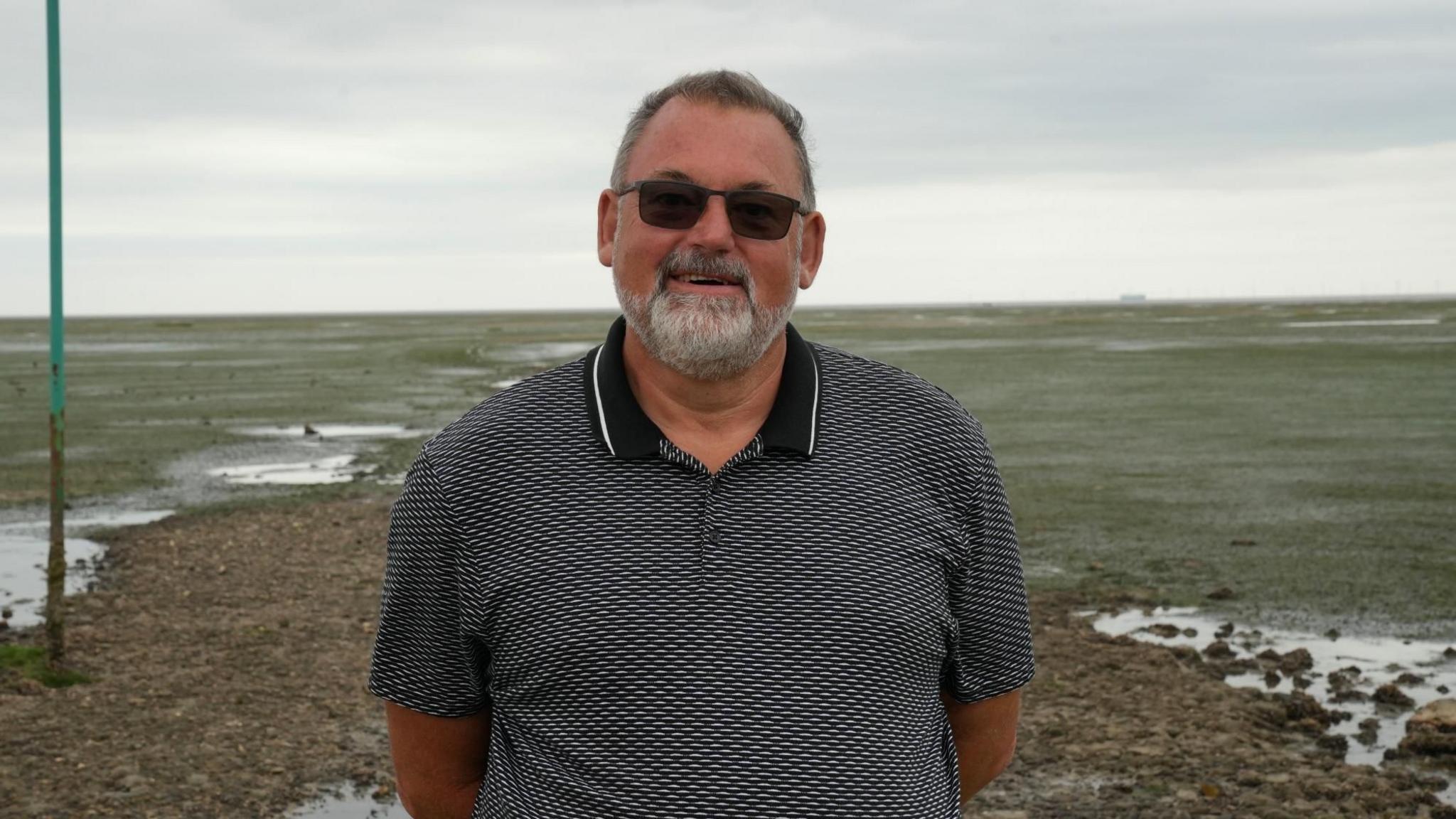 Kev Brown, a man with short, greyish hair and a grey goatee beard, is wearing sunglasses and smiling. He is wearing a polo shirt with a black collar and a grey, white and black pattern. The scene behind him is large, flat, empty of people, and looks like a green-ish brown coastal landscape.