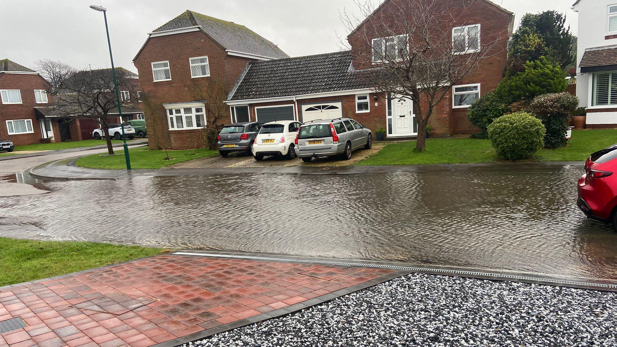 Flood water around a foot deep covering the entire road and up to the pavement. A red car is sitting in the flood water and some cars parked on driveways are unable to get out. A semi-detached house can be seen in the background 