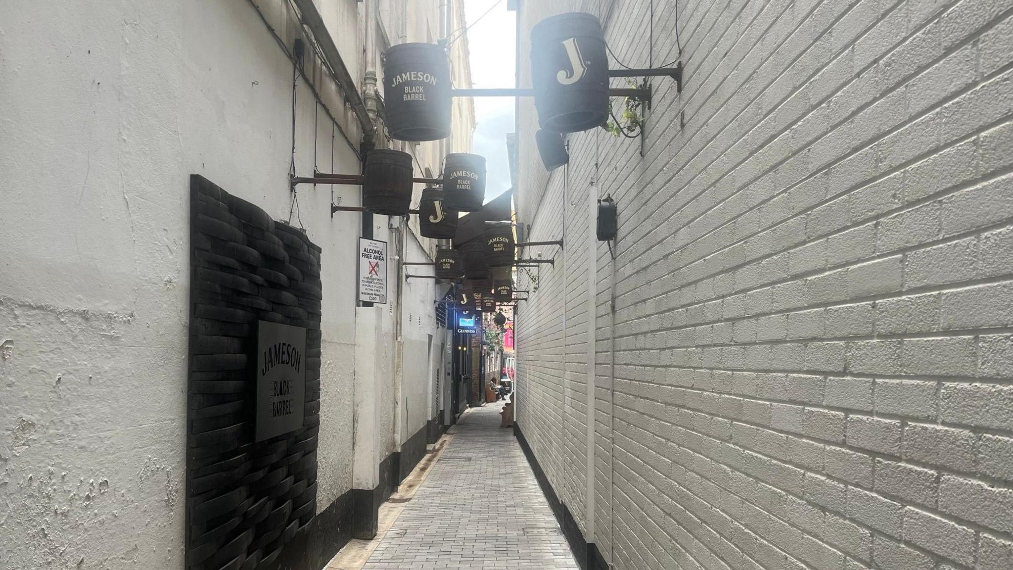 A narrow alley way with white walls and a white floor. There are brown barrels sticking out from high up