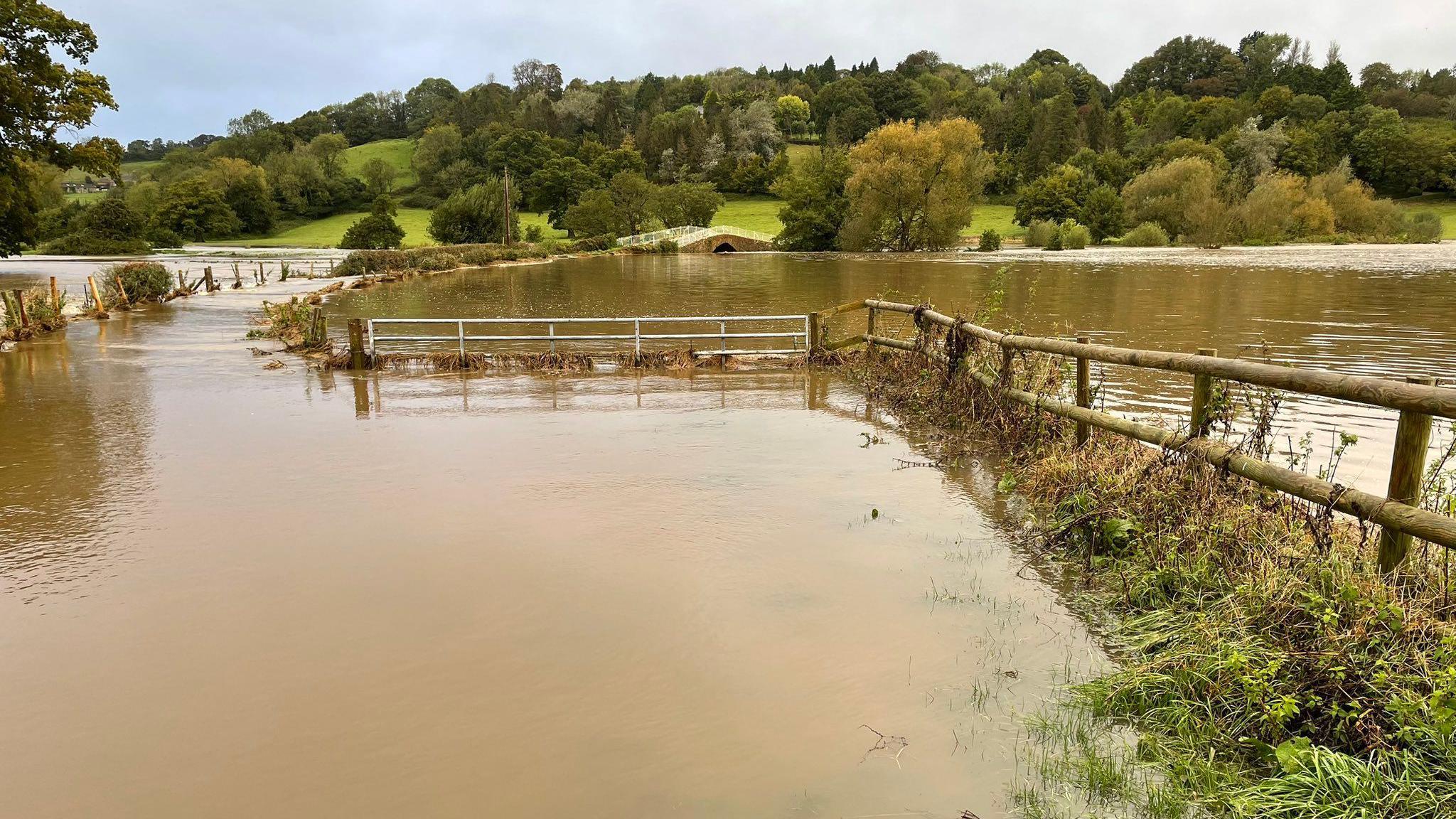 Image shows flood water covering a number of fields in Axminster, Devon. The water is covering grass and other foliage. A wooden fence can be seen separating the fields. Green fields and green trees are on a hill in a distance. There is a small bridge. 