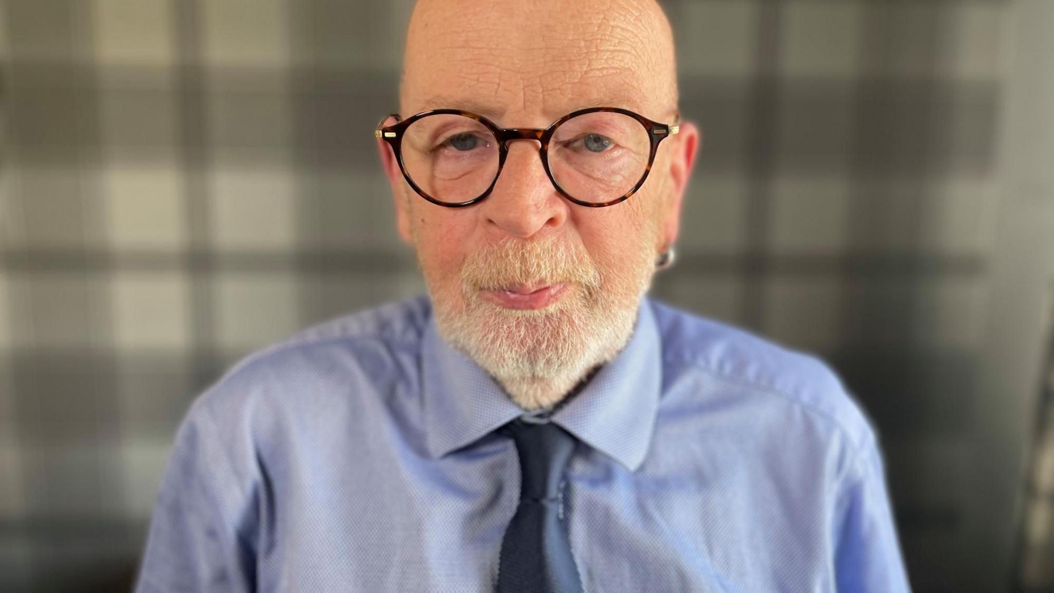 Stuart Hunter is photographed looking directly at the camera. He is a bald man with large round-rimmed glasses and a white beard. He is wearing a blue shirt and dark blue tie.