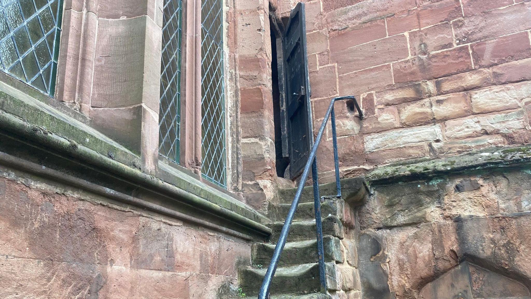 Old stone steps that are grey and green in colour with a black metal railing, leading up to a black wooden door in the side of a church.