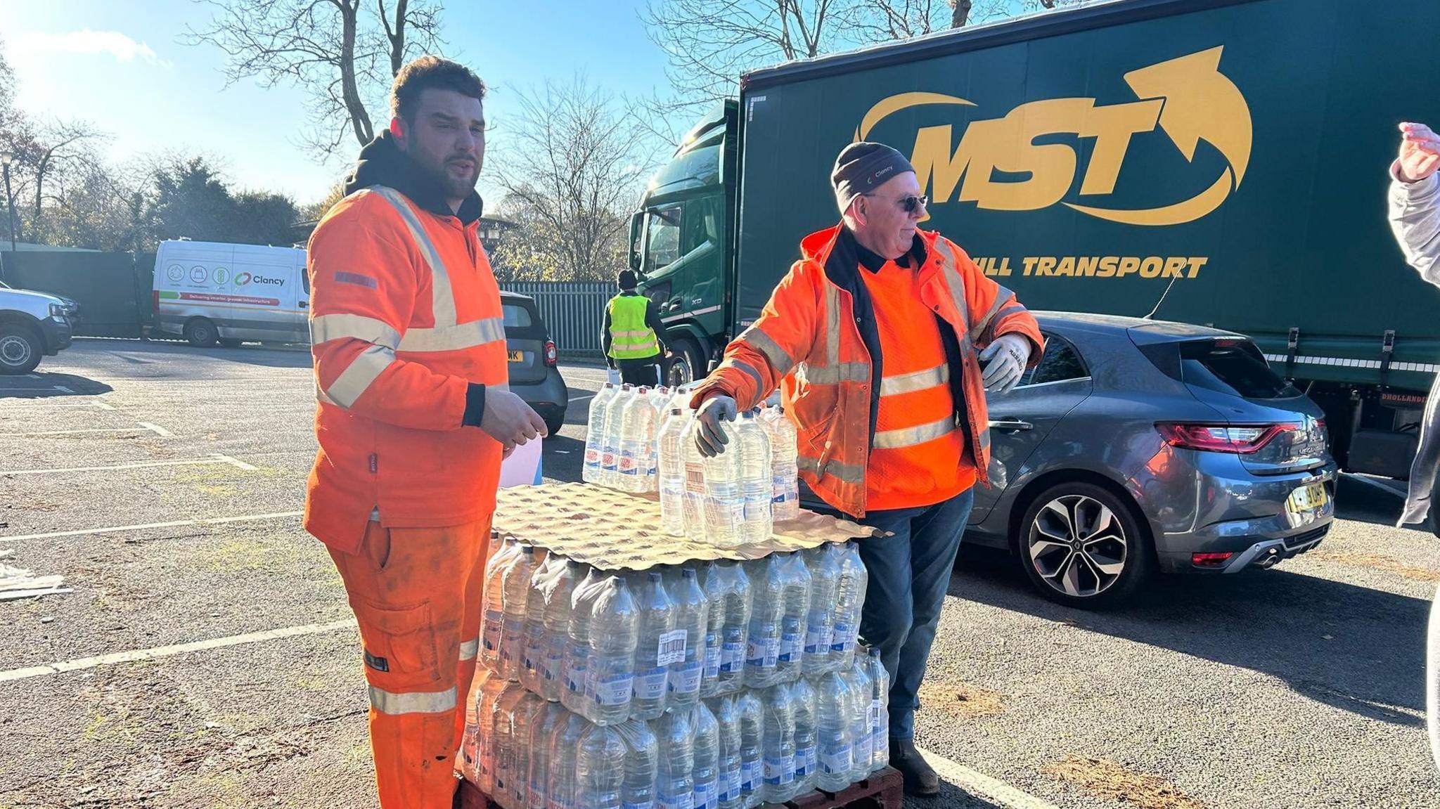 South East Water crew members at Tunbridge Wells Sports Centre on St John's Road, Tunbridge Wells. A temporary water station has been set up there. Both crew members are wearing orange high vis and are stood beside two crates of water bottles. 