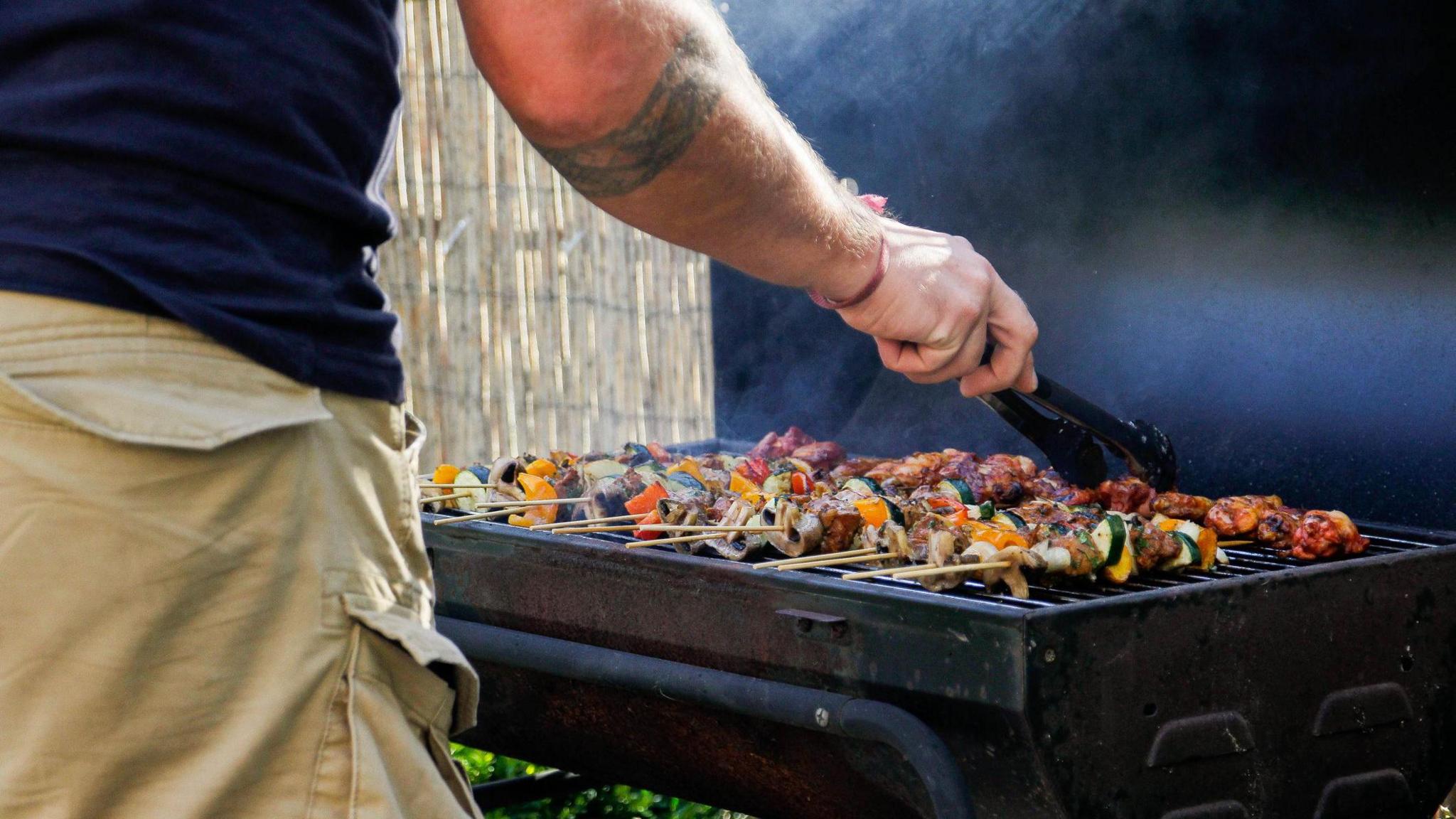 A close up of a man's arm holding a pair of tongs as he cooks meat and vegetable kebabs on a black barbecue.