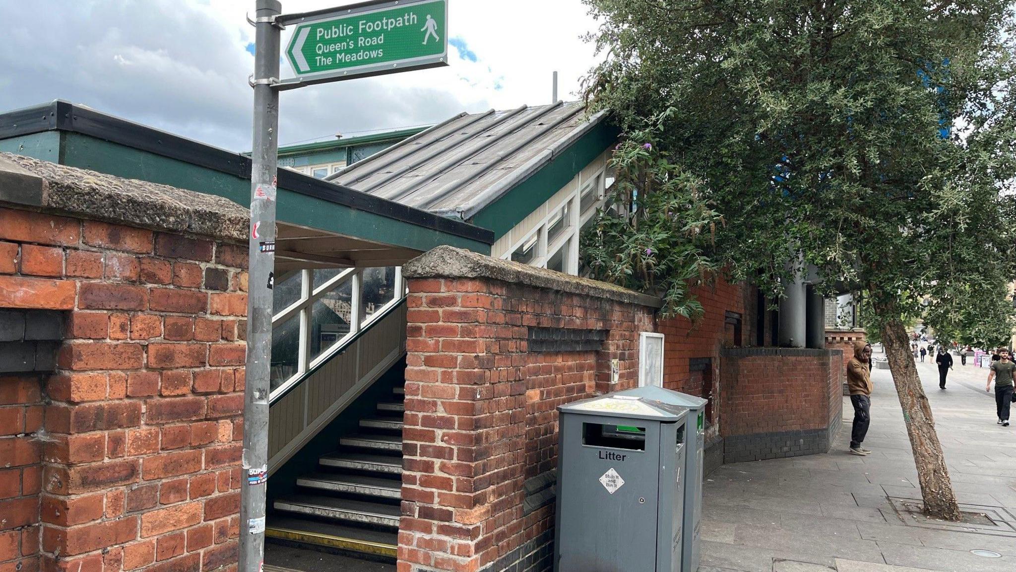 A street entrance to Nottingham station featuring a gap in a brick wall, a footpath sign and steps leading up into a covered walkway