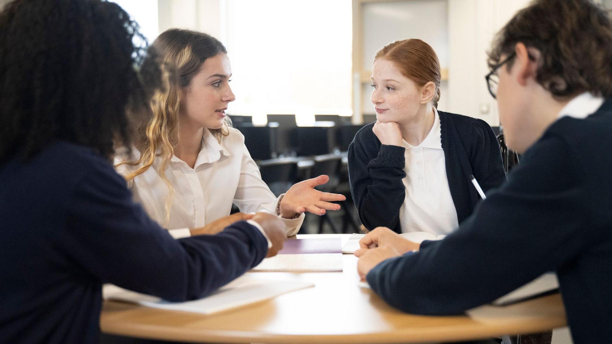 A stock image group of young people in a classroom sit around a table during a discussion. The two who have their faces to the camera are female and wear a navy and white school uniform. 