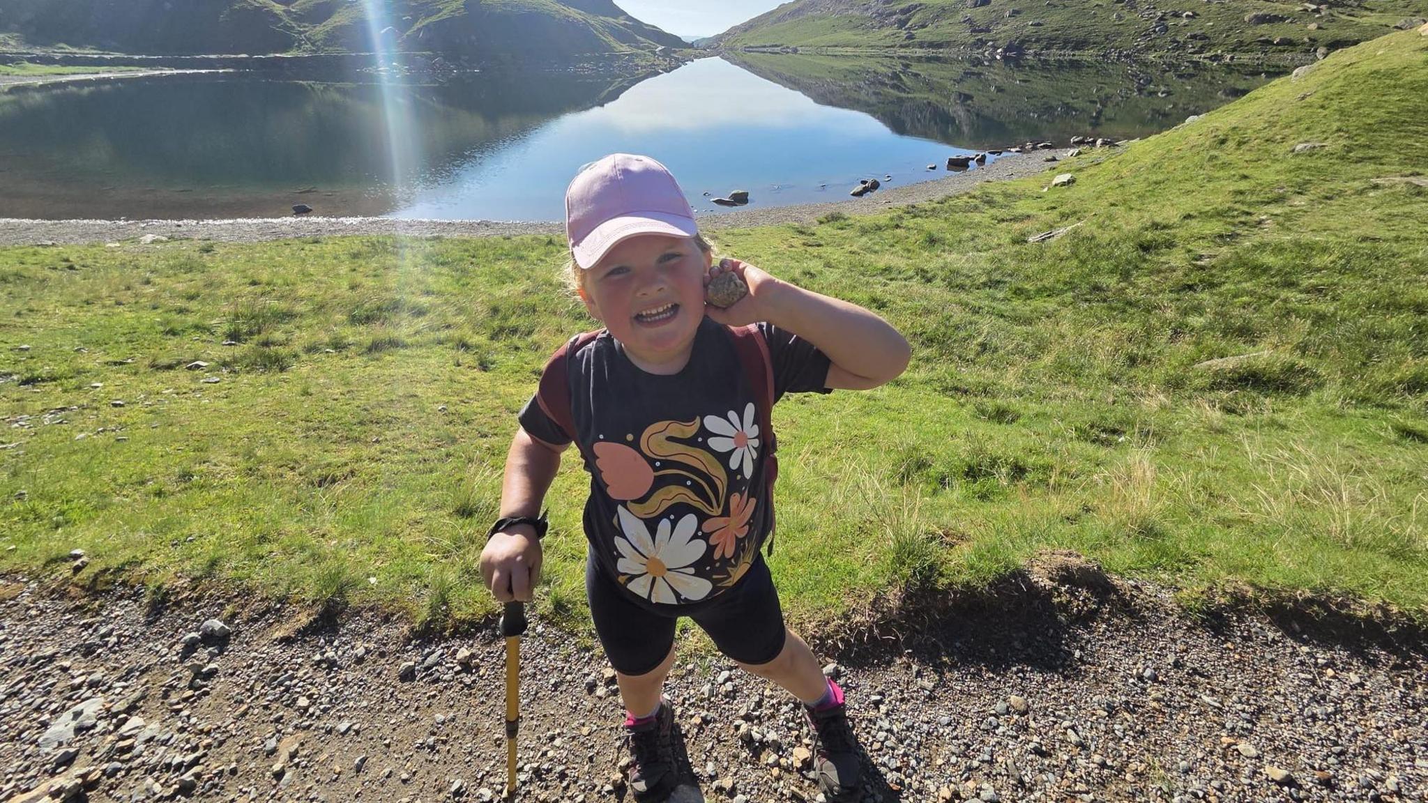 A six-year-old girl wearing a navy blue t-shirt, shorts and a pink baseball cap. She is stood on a footpath with an area of grass and a lake behind her.