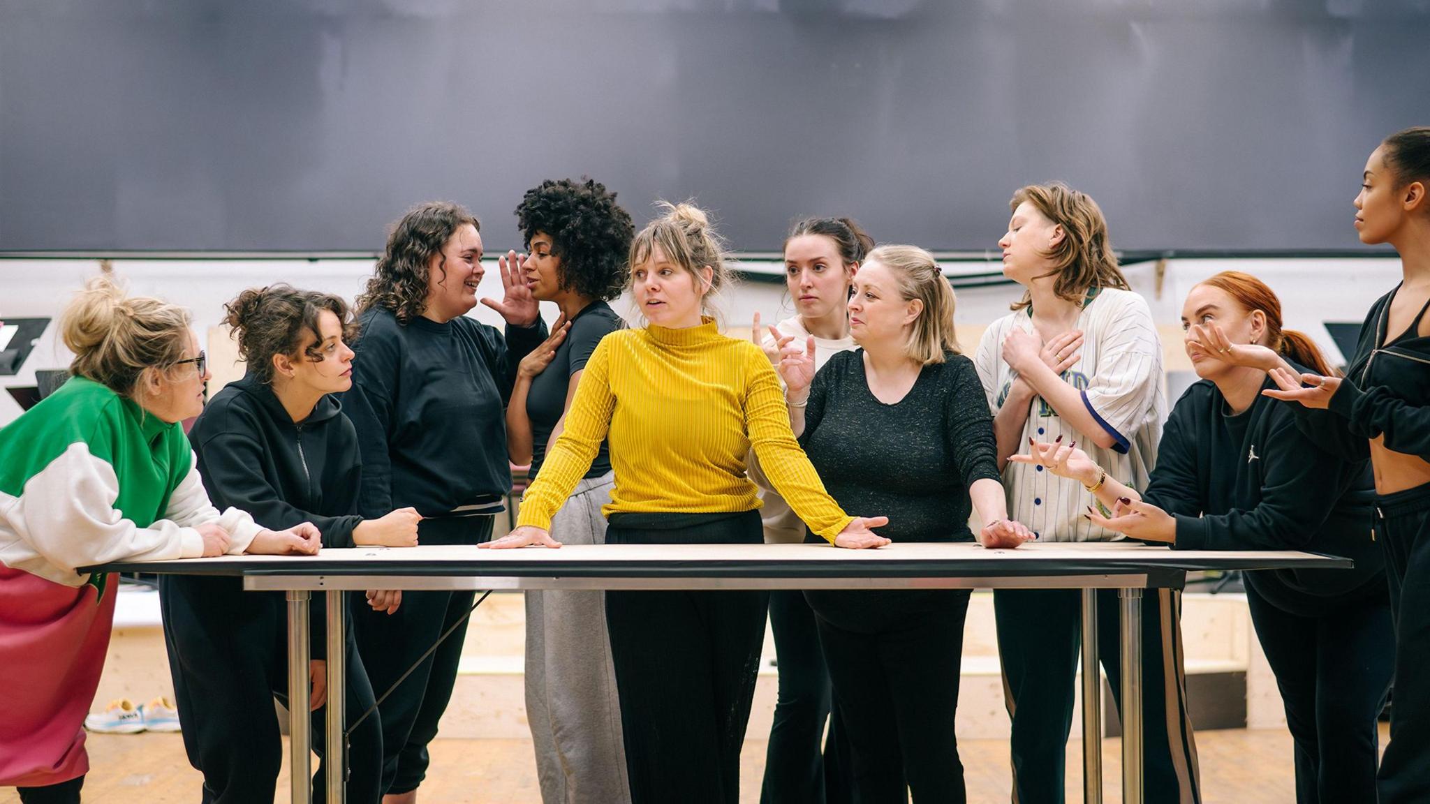 Dawn Sievewright, centre in a yellow jumper and black trousers, rehearsing for her part in Wild Rose surrounded by nine other performers standing around a large table.