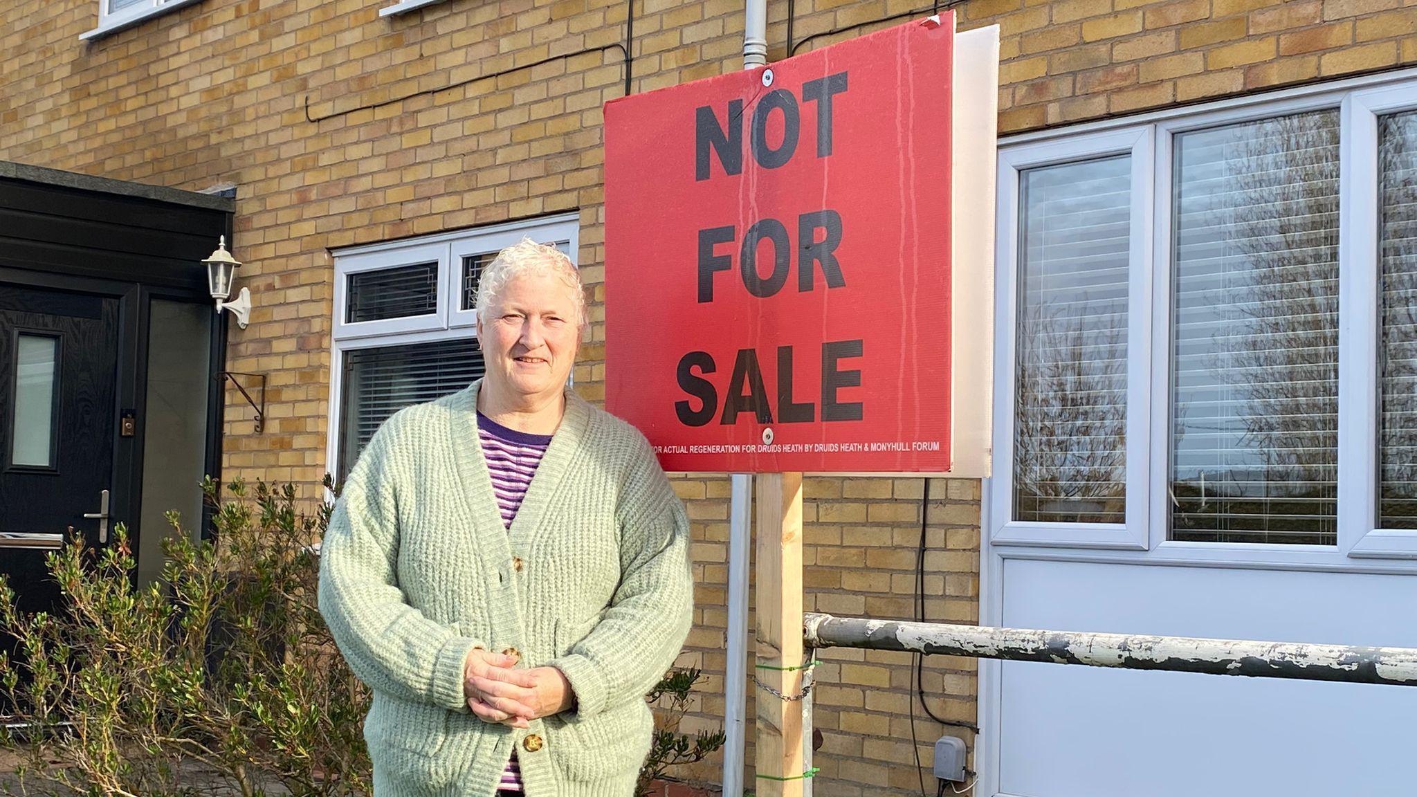 A woman with short white hair is wearing a pink, purple and white striped T-shirt with a sage green cardigan on top. She is standing outside a house with a large red sign. In black writing it reads "not for sale"