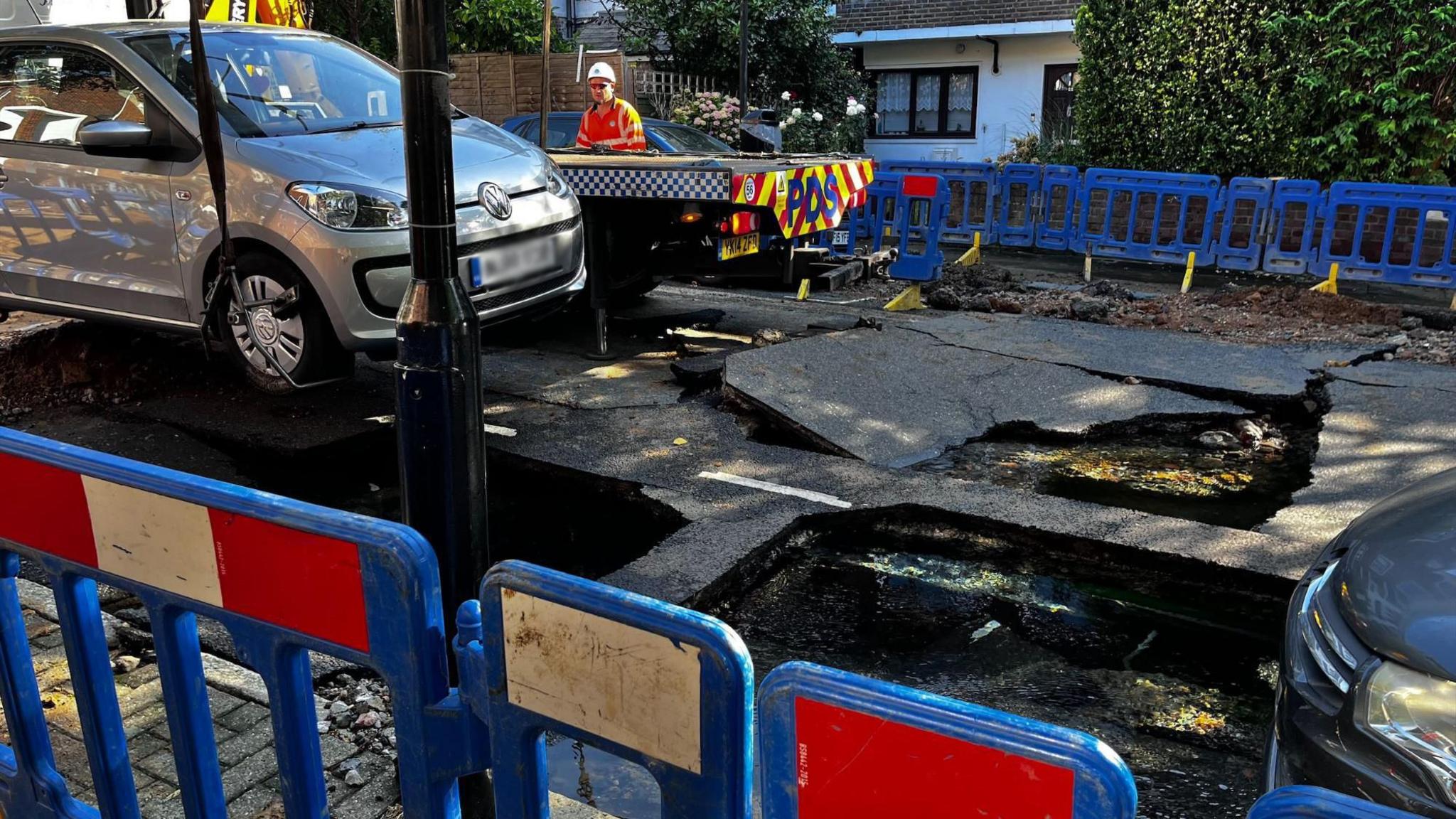 A road that has been broken up with large holes in it. There are blue barriers with red and white markings on either side of the road and a recovery truck on the far side. A small silver Volkswagen car is being lifted off the road.