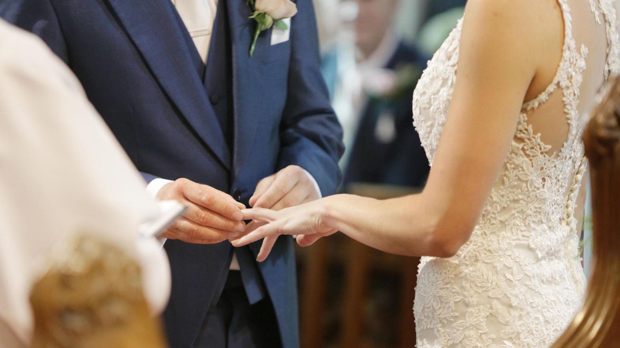 A person, wearing a blue suit with a white flower attached to the chest, placing a wedding ring on a woman's outstretched hand. She is wearing a wedding dress.