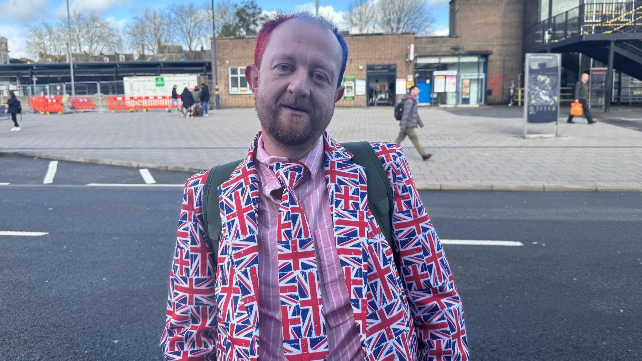 Bartly is dressed in a salmon pink shirt with white stripes, and a British flag tie and jacket. He has dyed pink/purple hair and a brown goatee beard. He has rucksack straps over both shoulders and is standing with the station in the background.