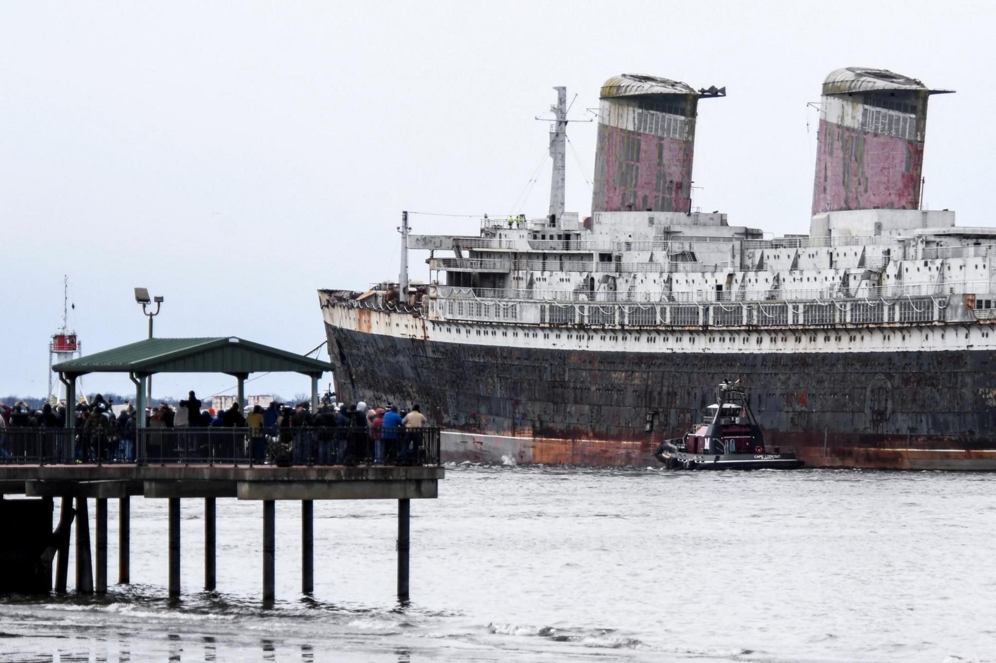 Onlookers watch the SS United States as she is towed out to sea by tugs. The ship looks old and rusty.
