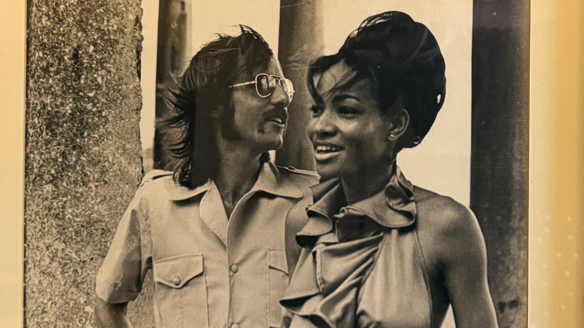 Dave Swaffer in a black and white photograph wearing a shift and sunglasses with long hair. He is with Judith who is wearing an elegant dress and has her hair styled so that it is up.