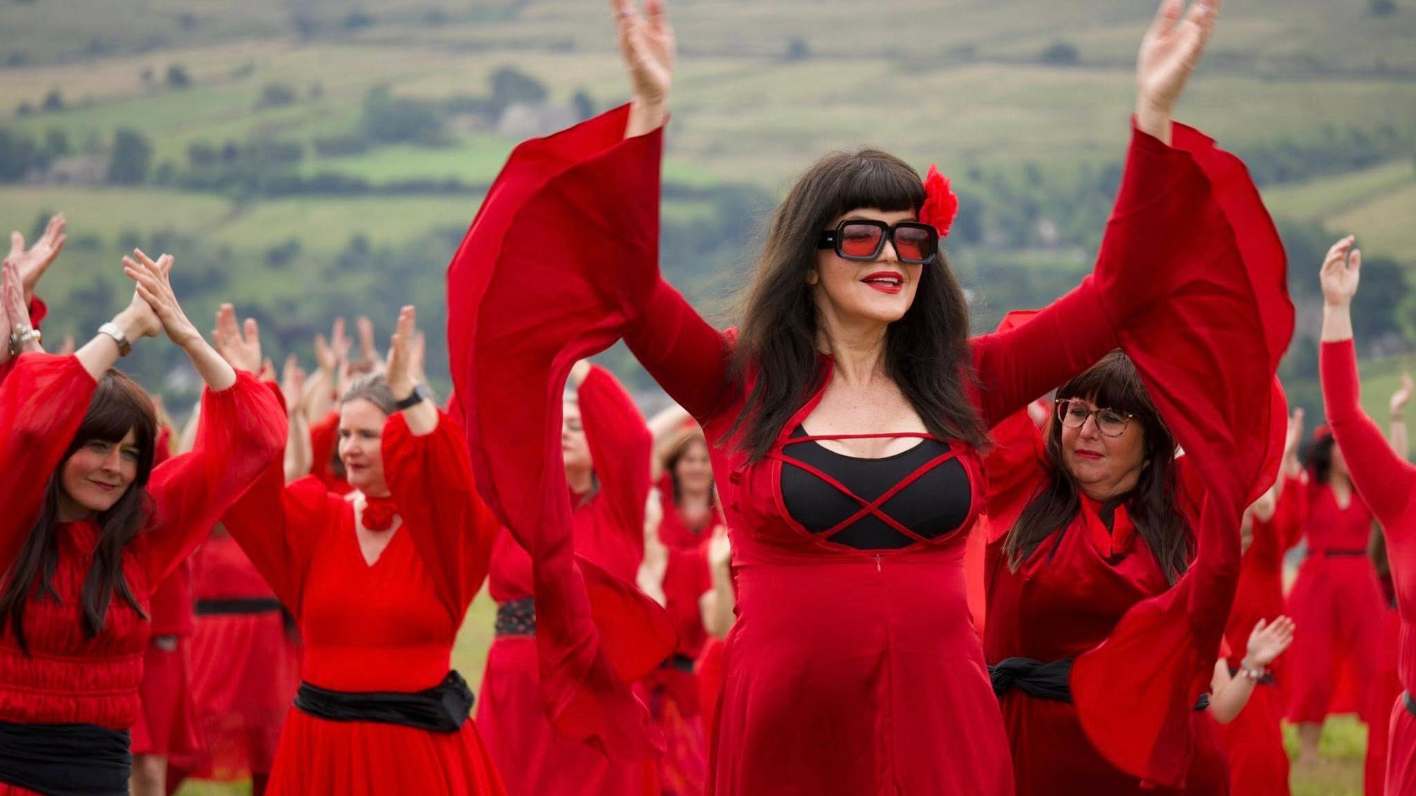A group of women in red dresses dancing on the moors - the one in front has long brown hair, sunglasses and a flower in her hair.