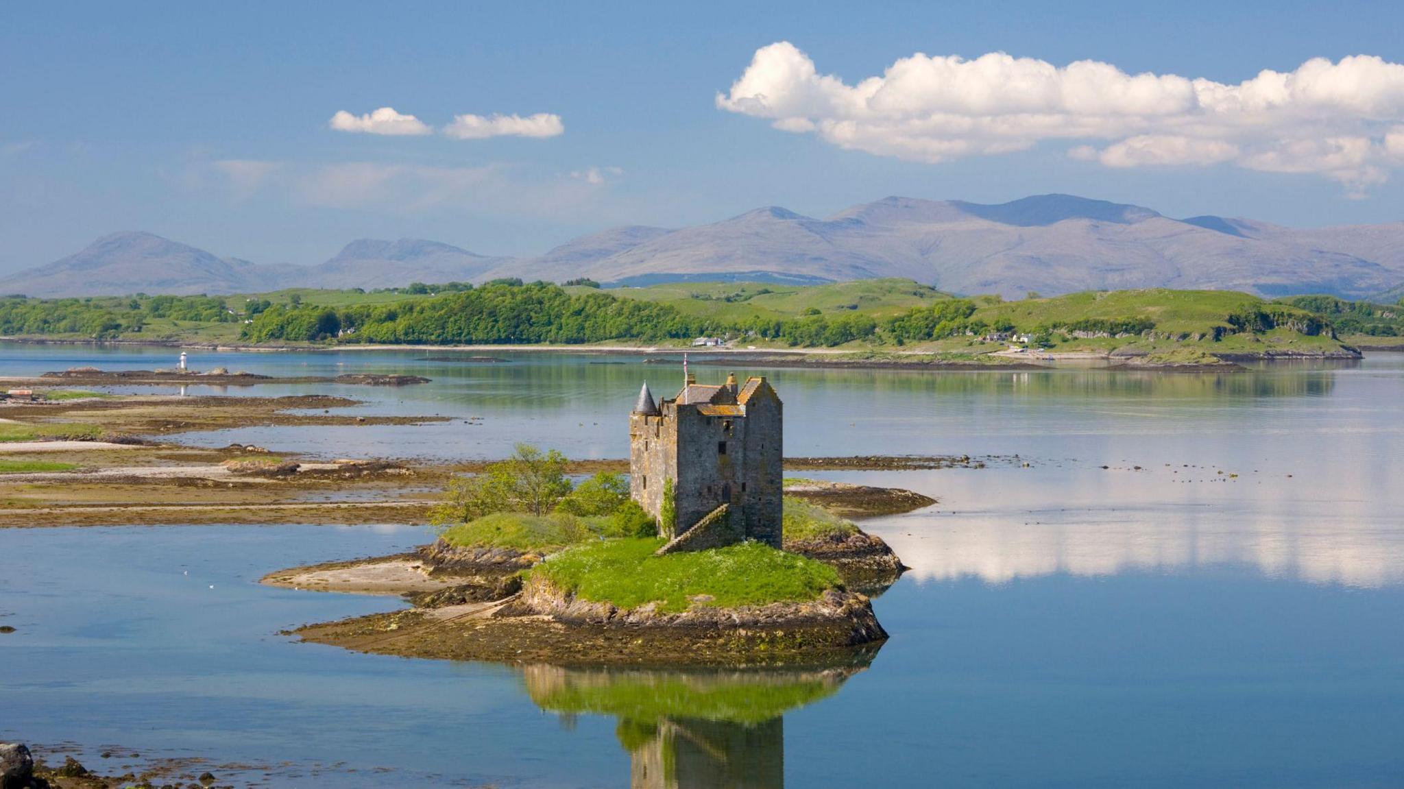 Seann chaisteal cloiche, le tùr, agus pòla brataich, air eilean beaga ann an loch-mhara. Tha eilean nas motha le craobhan air am meadhan an deilbh agus beanntann air cùl sin fada às. Chithear faileas dhen chaisteal agus na sgòthan san uisge.