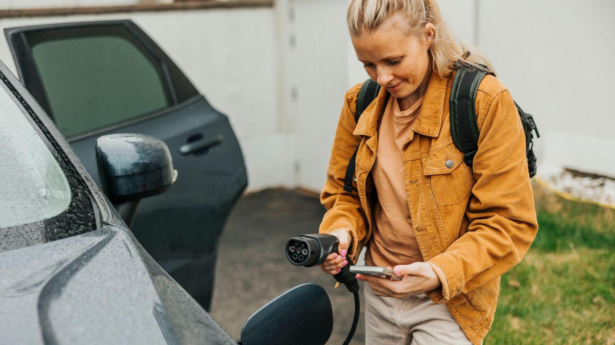 A young woman with her phone in her hand, about to plug an electrical charger into her car - she is wearing an orange jacket and has her blonde hair tied back into a pony tail, and is standing next to the car with the door opened