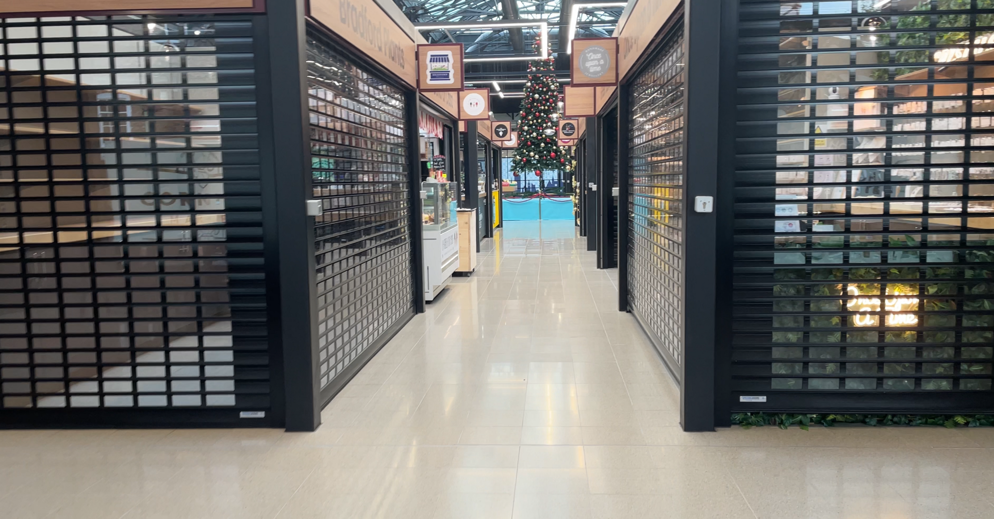 A view of stalls at the Cornhill Market in Lincoln. Several have black shutters down to show they are closed and the walkway is also empty. A Christmas tree is visible in the distance.