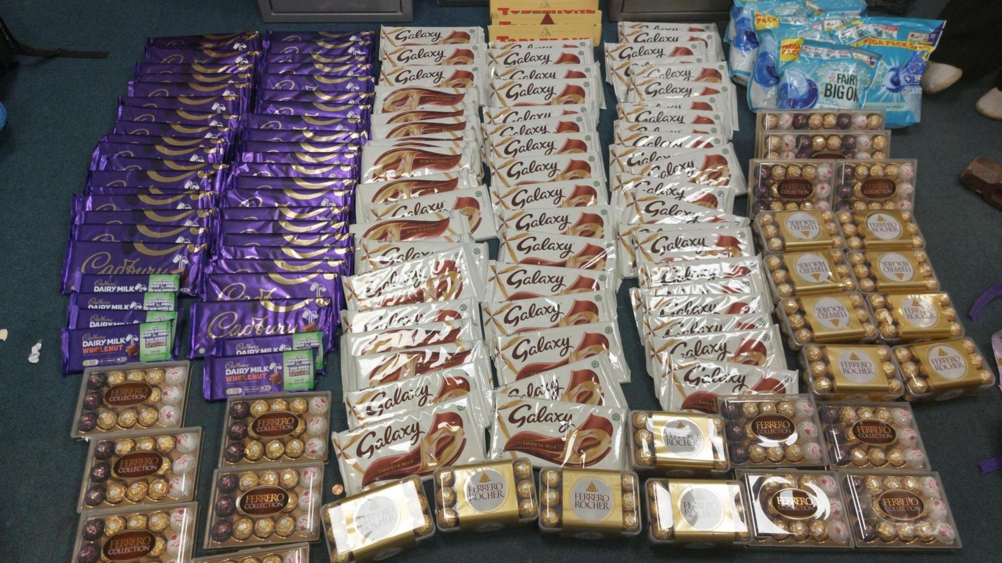 Lots of bars of chocolate and boxes of chocolates are lined up on a dark background. There are also some bags of washing tablets.