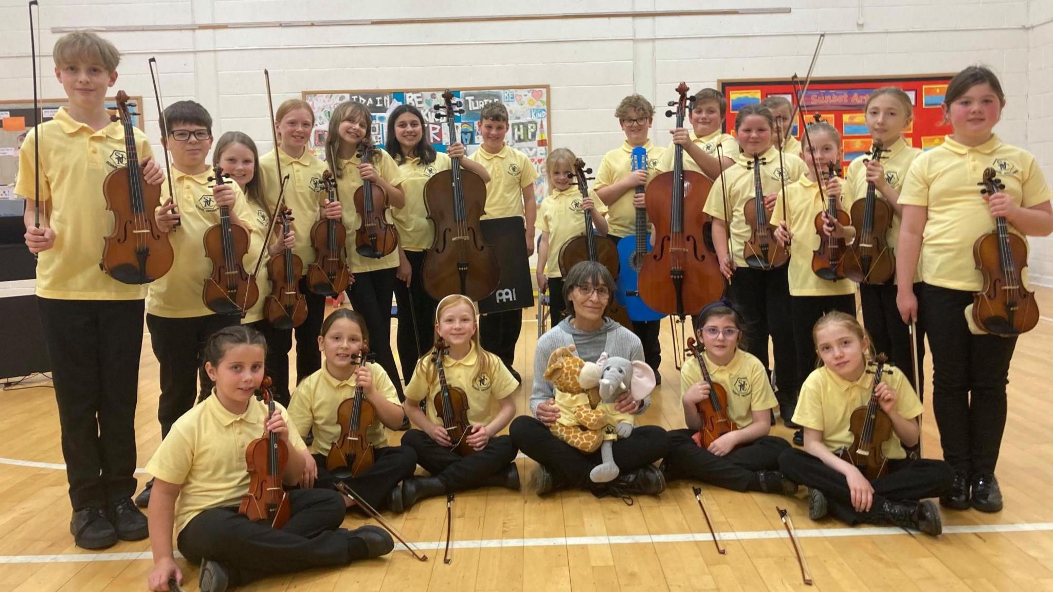 A large group of young children in matching yellow polo shirts smile holding up violins