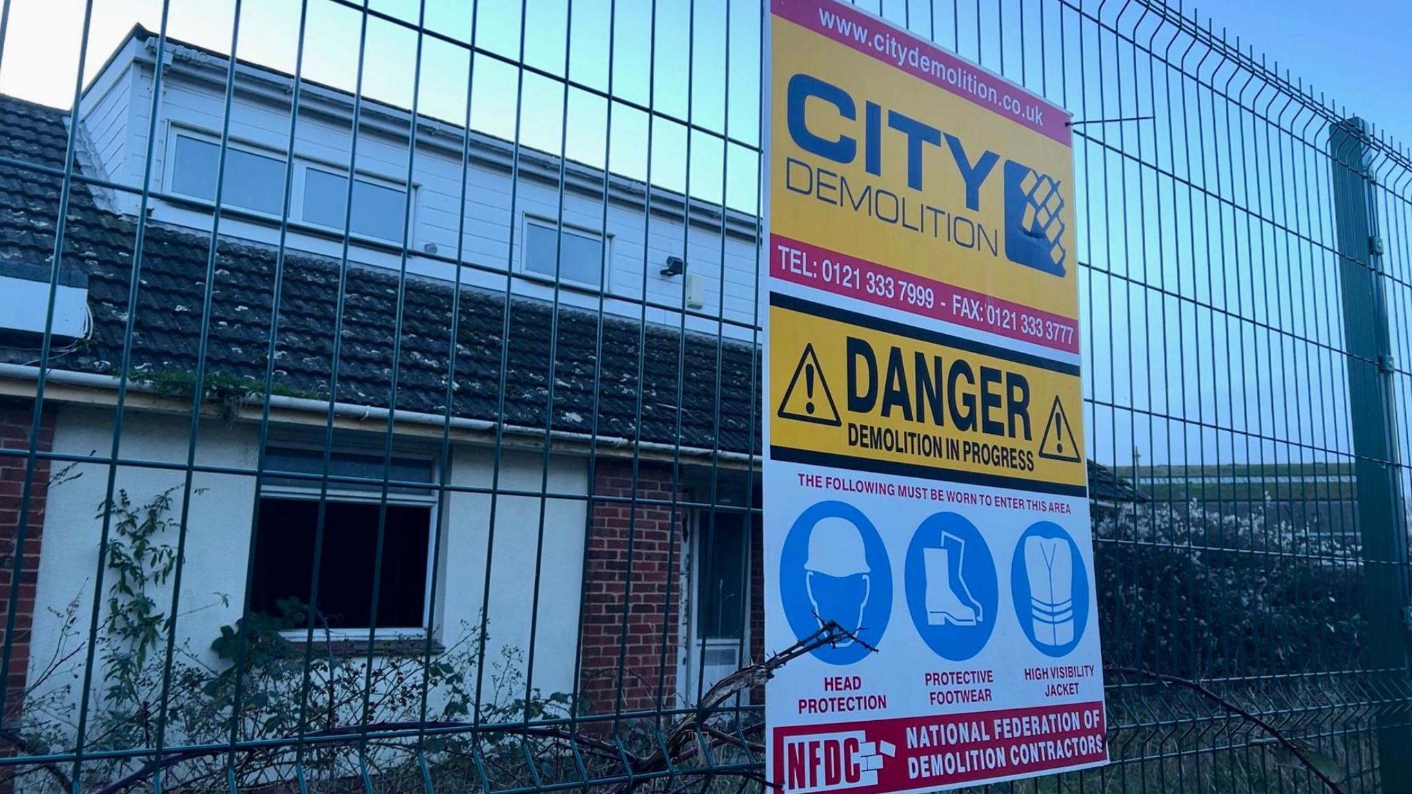 A demolition sign attached to a fence in front of a derelict house on the Water Lane site