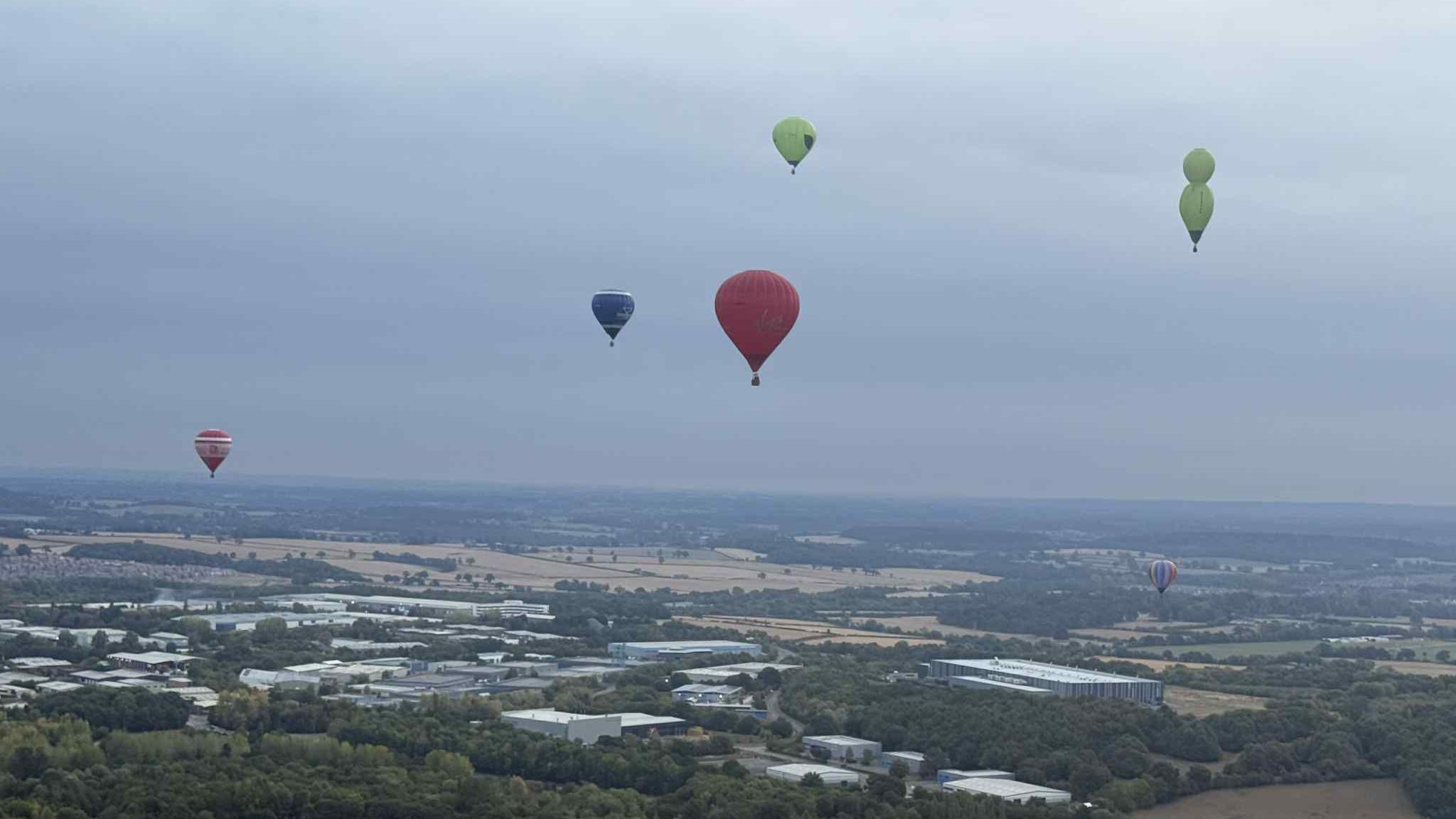 View of multi-coloured hot air balloons in the sky while trees, a car park and buildings are below. The skyline looks grey due to the weather.