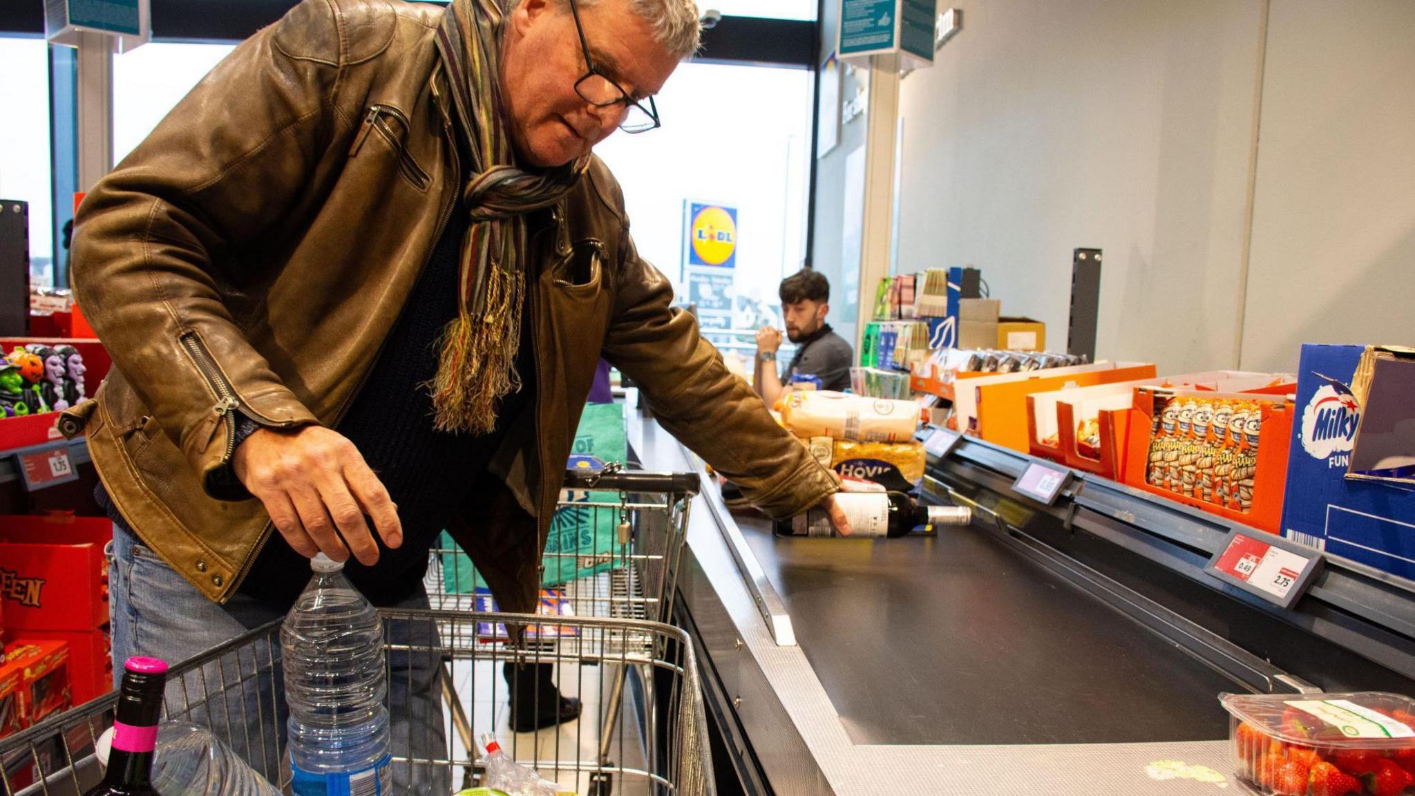 A middle aged man wearing a brown leather jacket and striped scarf loading a trolley of shopping on to the conveyor belt at Lidl