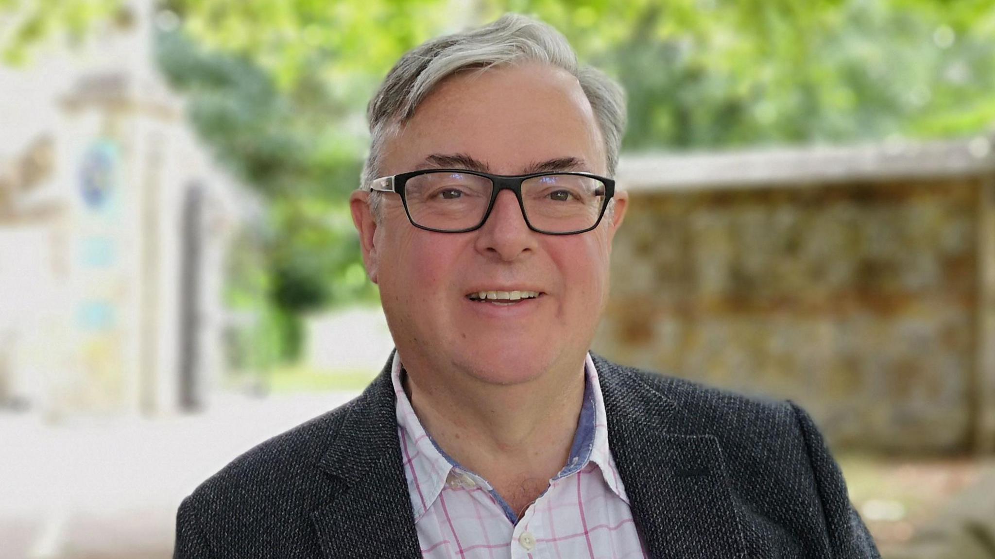 Gavin Grant is standing under a tree in a village square. He is wearing a grey jacket and white shirt. He is smiling at the camera.