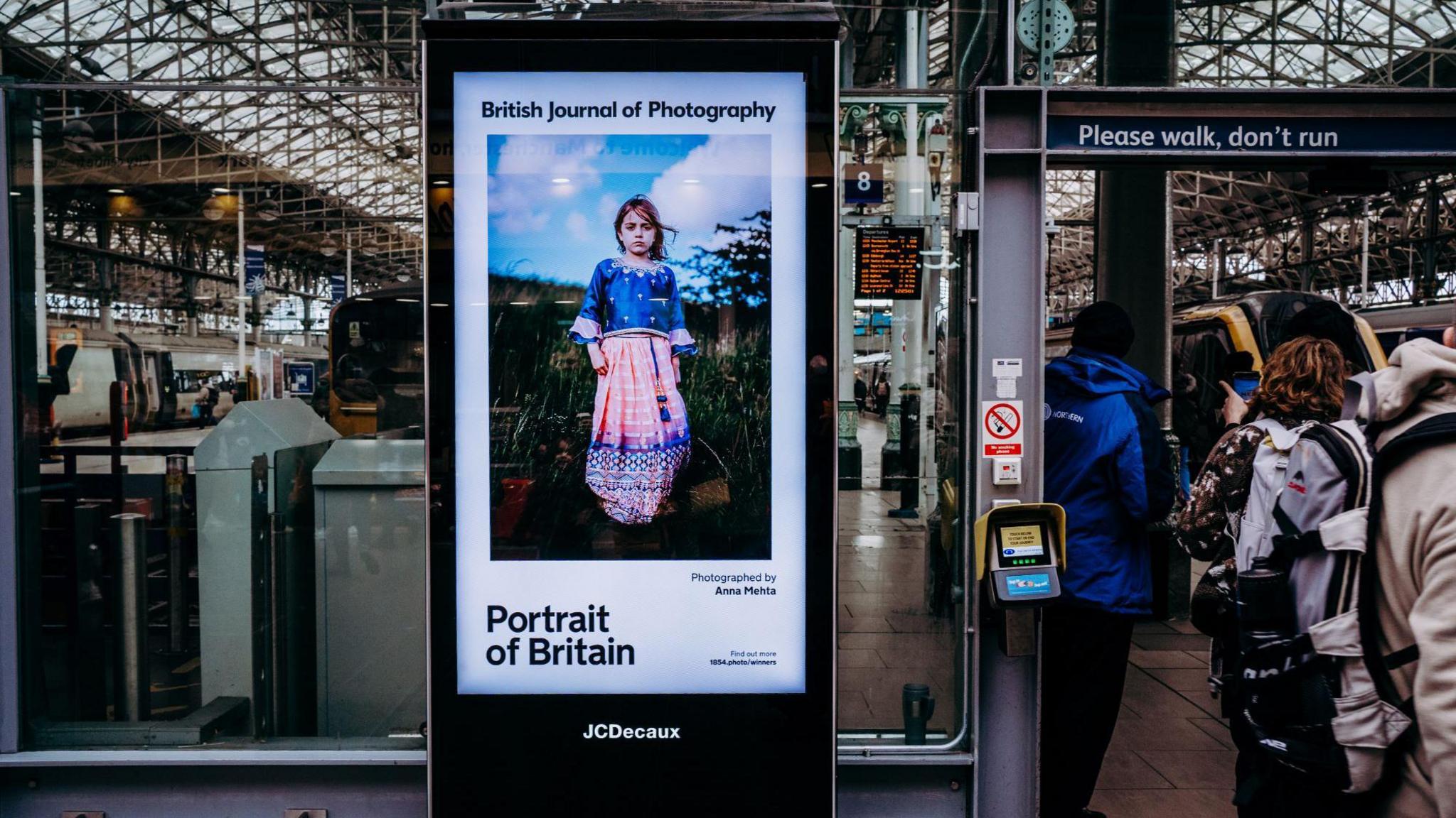 A portrait of a girl in an Indian dress standing in a rural area is shown on a train station advertising board.