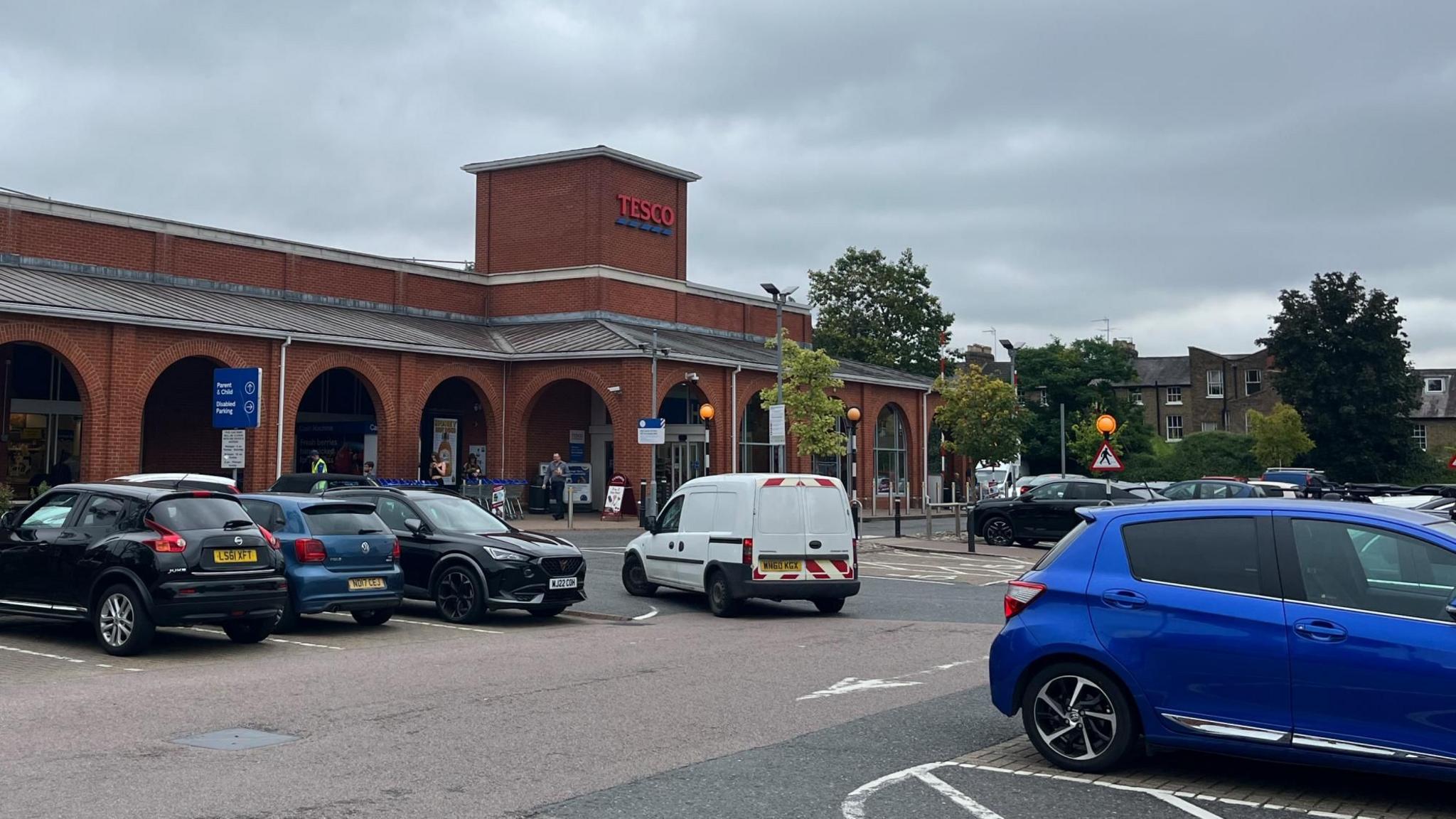 View of the front of red brick-fronted Tesco in Hertford, with vehicles parked and being driven in the car park 