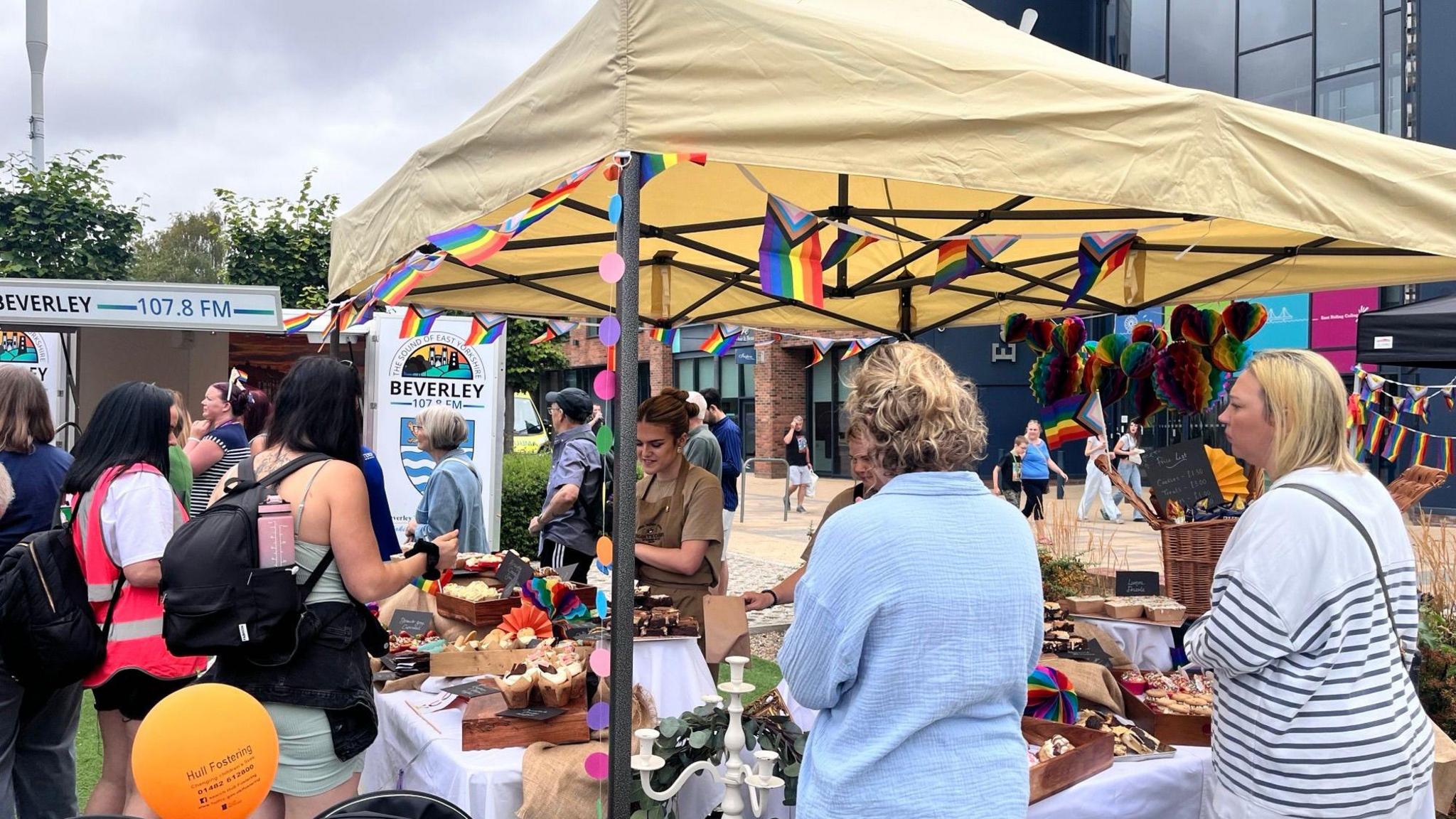 People gathering around a market stall which has a yellow canopy and is selling baked goods.