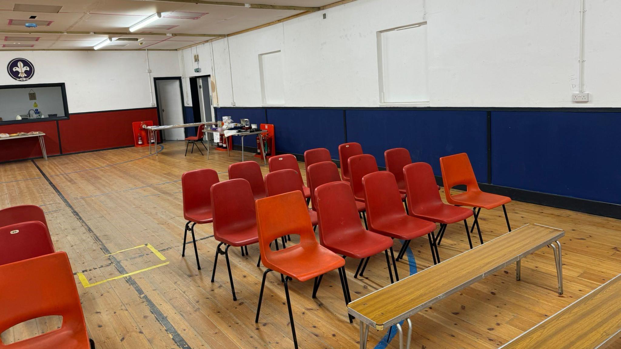 Several rows of orange chairs fill a hall with a couple of benches in front. The floor is pine and there's a hatch into the kitchen. 