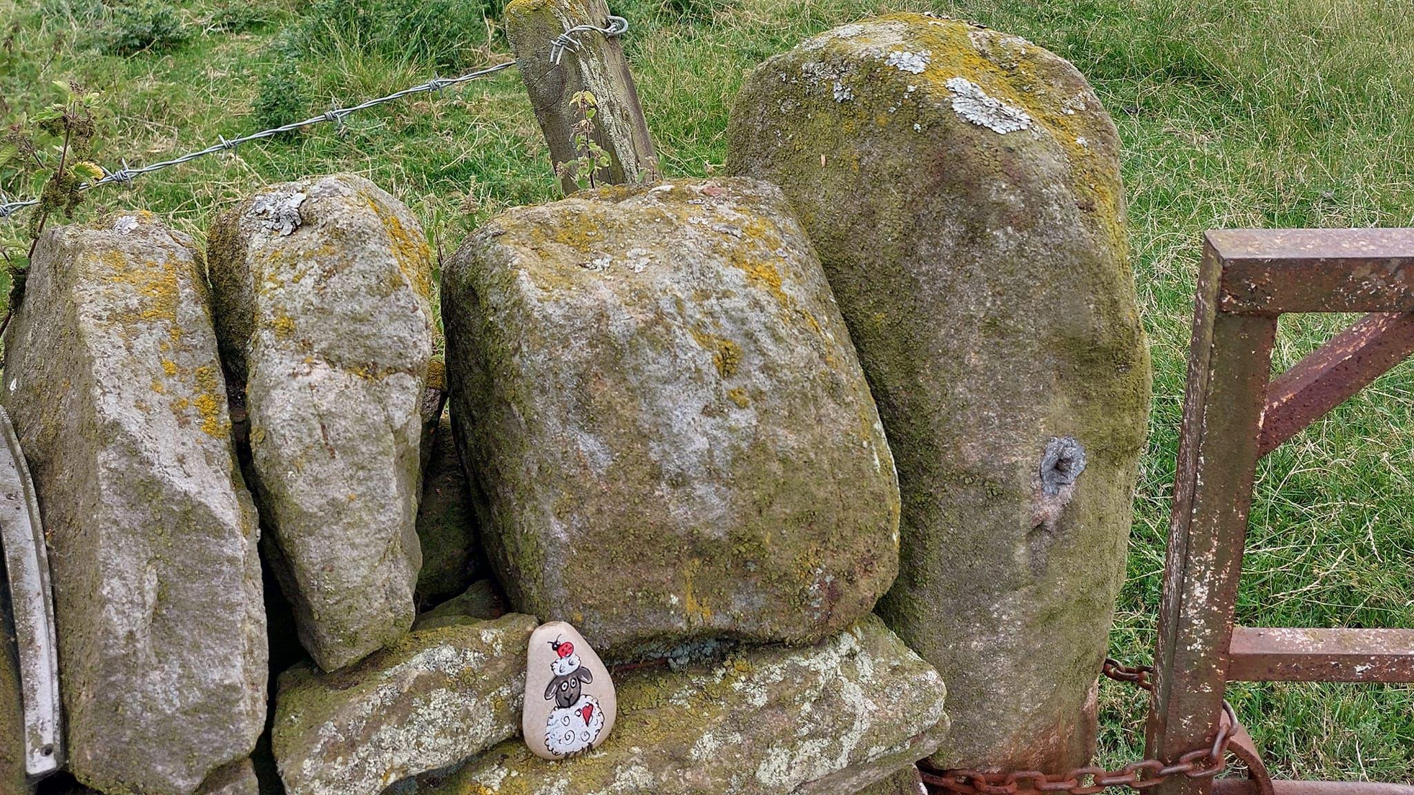 A small painted rock depicting a cartoon sheep is placed on a stonewall in a field.