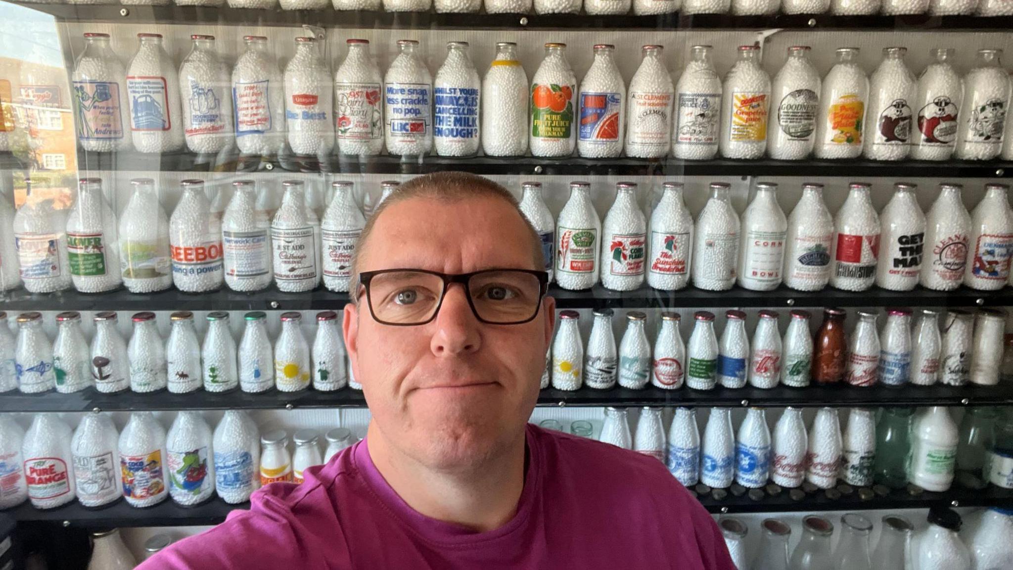 Paul Luke, who is wearing glasses, poses in front of milk bottles - all of which are stored behind glass